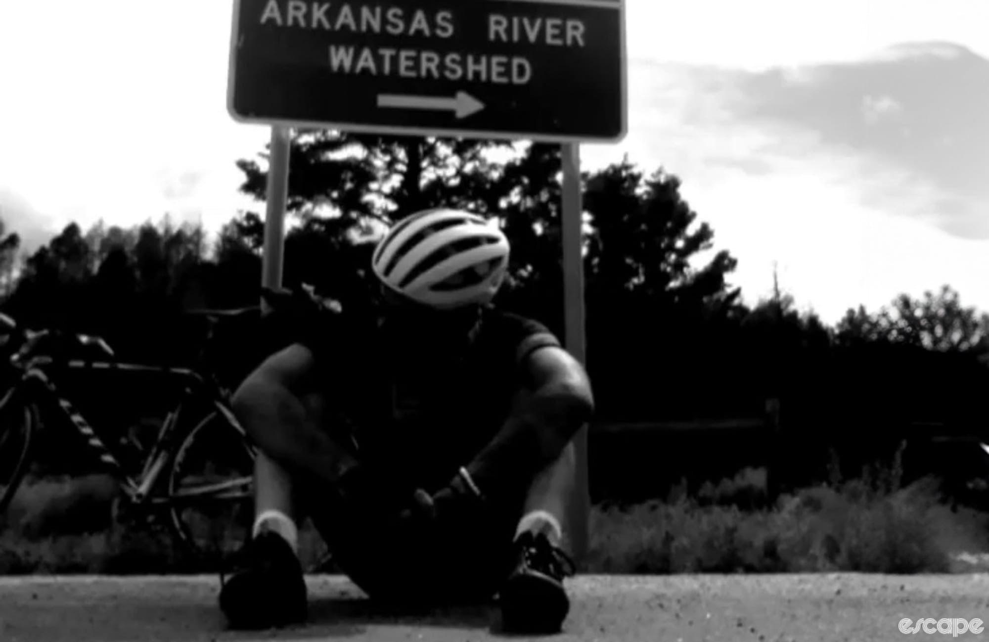 A cyclist sits exhausted on a road below a road sign, in a noir-style black and white photo.