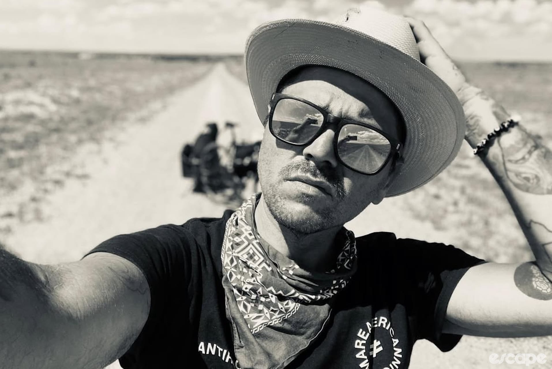 A man wearing sunglasses, a wide-brim hat and bandanna takes a selfie on a deserted road.