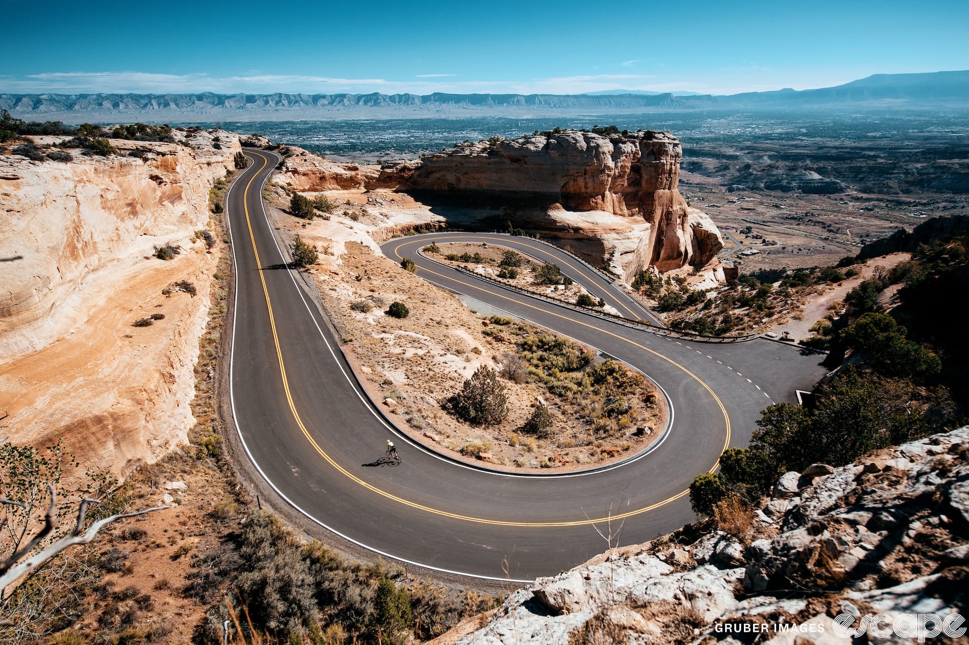 A lone cyclist is dwarfed by sweeping switchbacks ascending through a desert landscape.