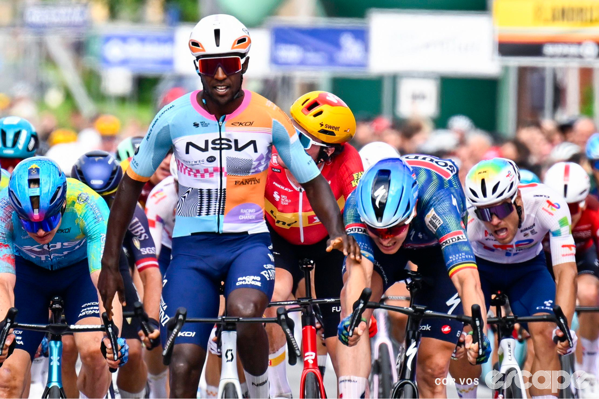Biniam Girmay, in his current NSN kit, begins to raise his arms in celebration ahead of a speeding peloton. 