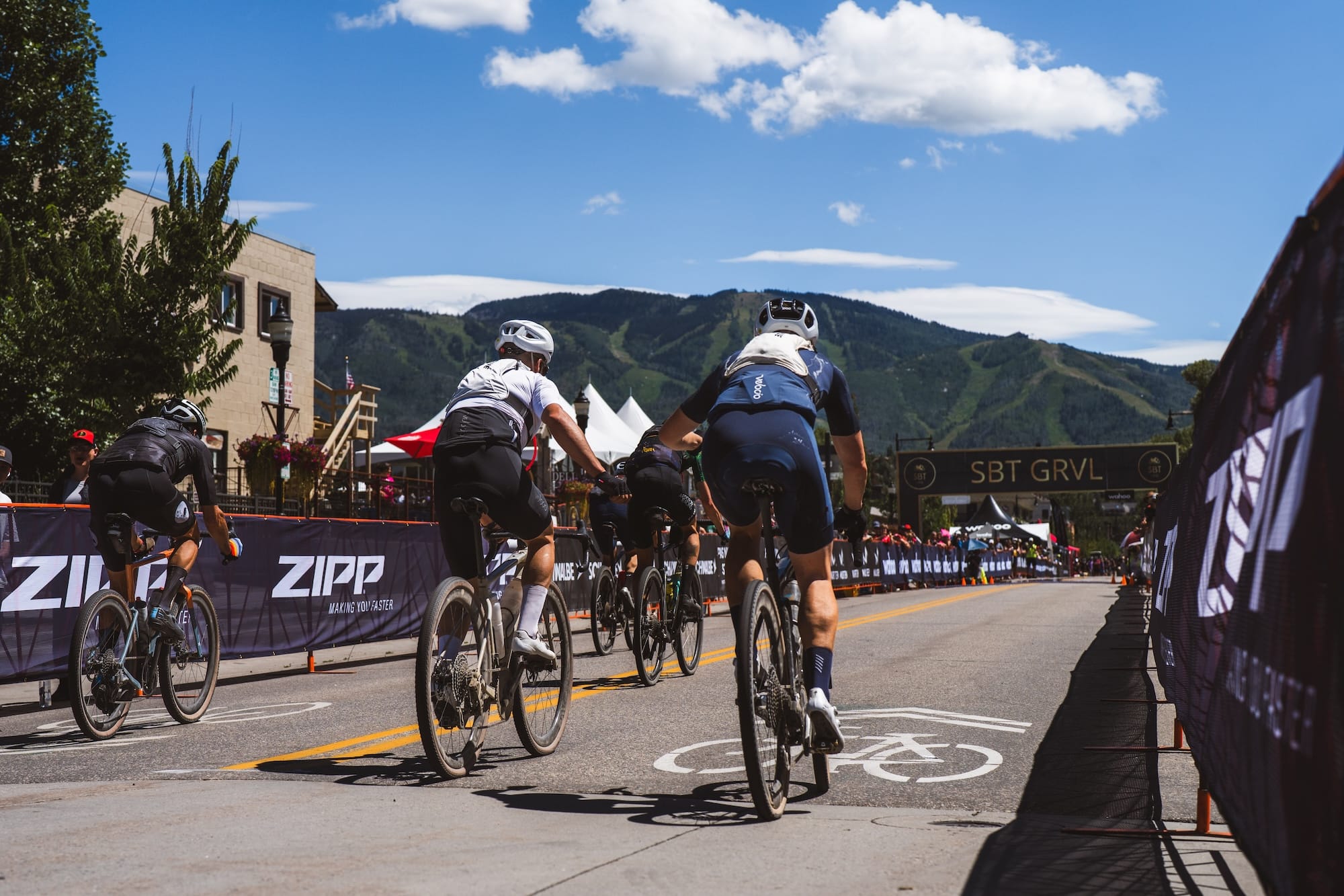 Two riders shake hands as they near the SBT GRVL finish arch in Steamboat, Colorado, with green mountains in the background.