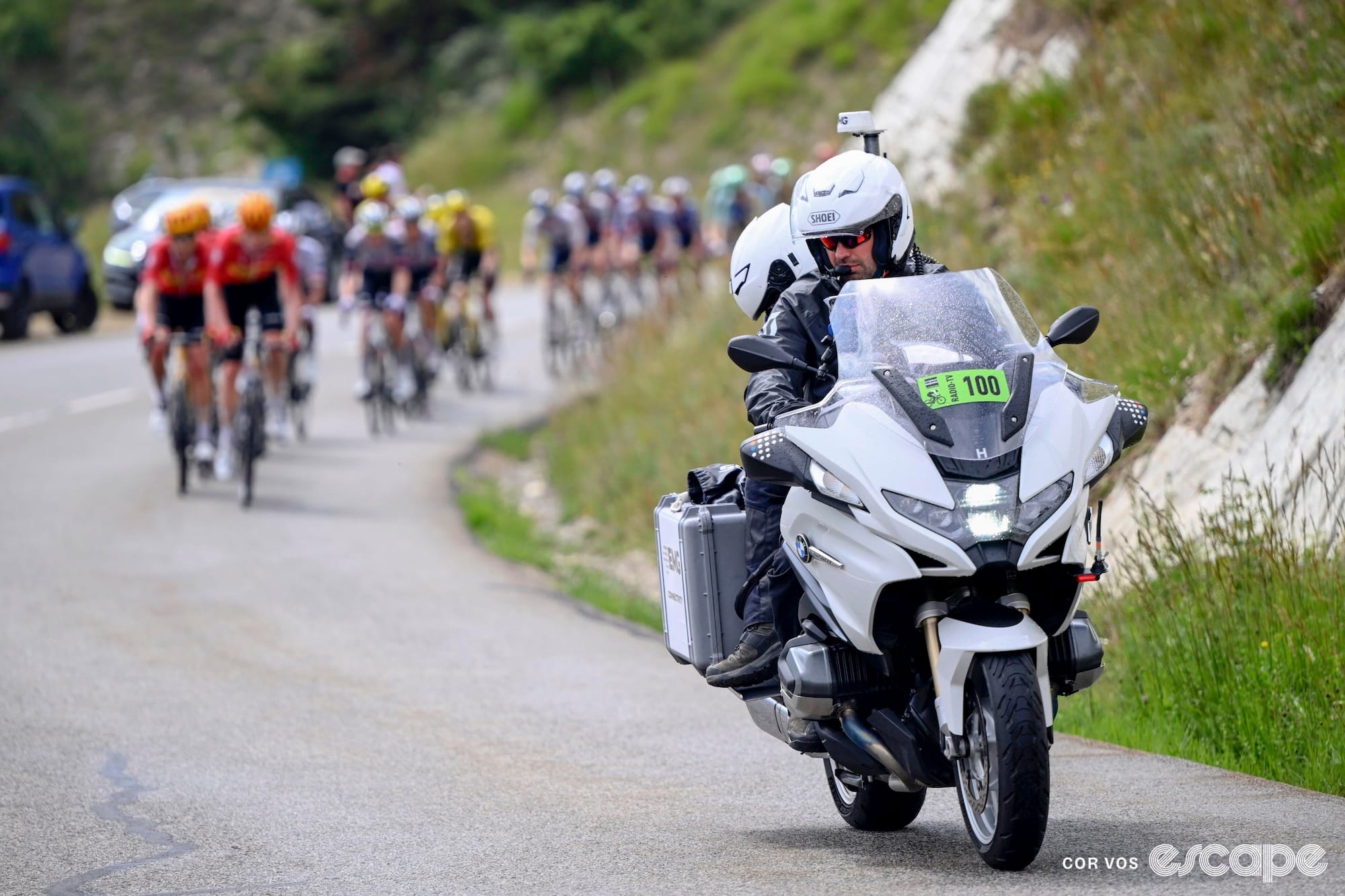 A television moto in the foreground rides ahead of the peloton up a winding mountain road.
