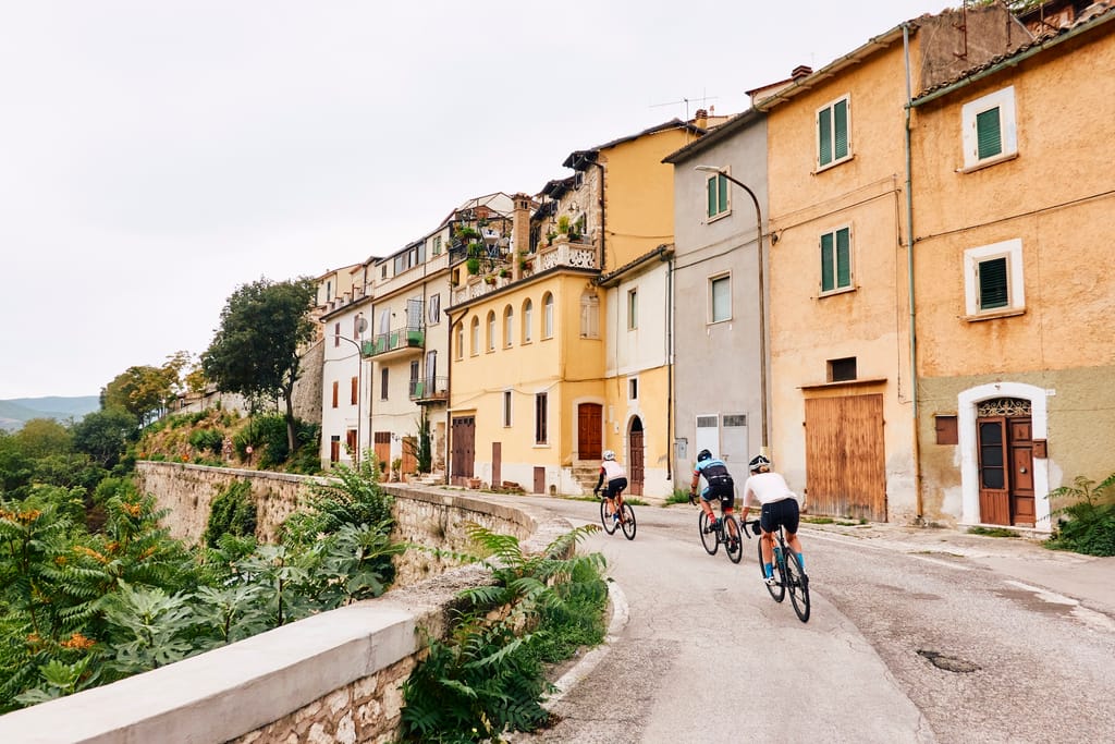 A group of three riders passes through an Italian hilltop town, with a row of houses to the right and a stone retaining wall and hill to the left.