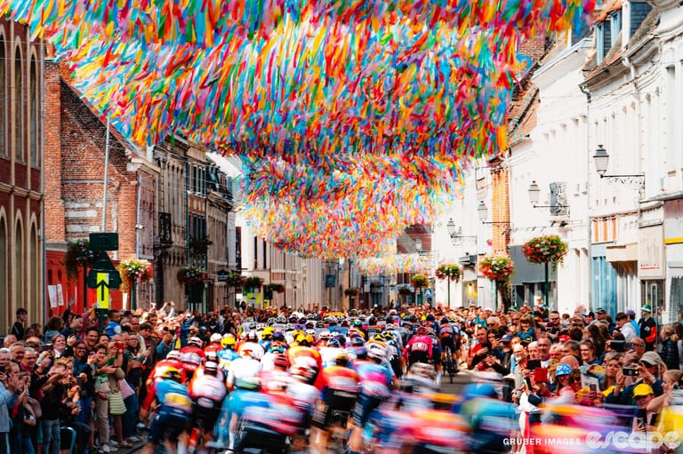 A blurred mass of riders races down a narrow street at the Tour de France under a swirl of colorful ribbons strung between buildings.