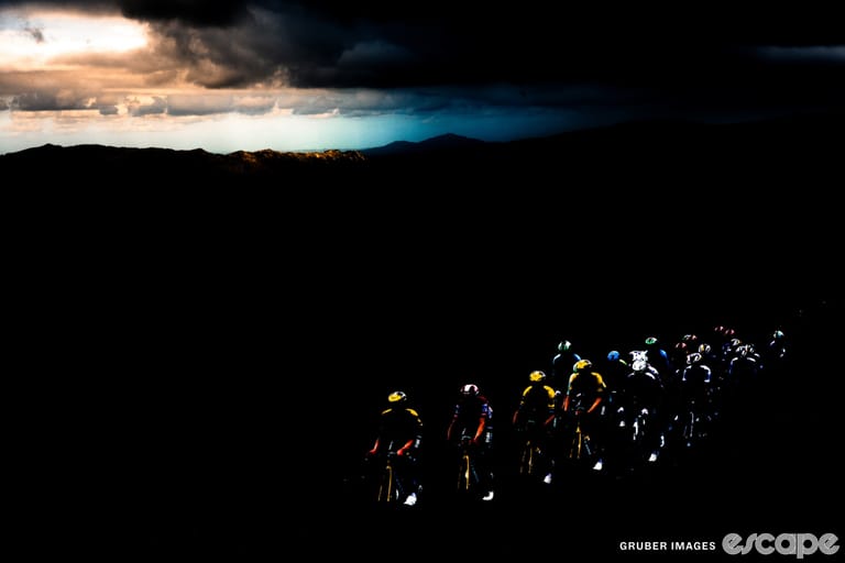 A peloton of riders climbs against a mountain backdrop. The shot is darkly lit, emphasizing a dramatic, almost black background with pops of color.