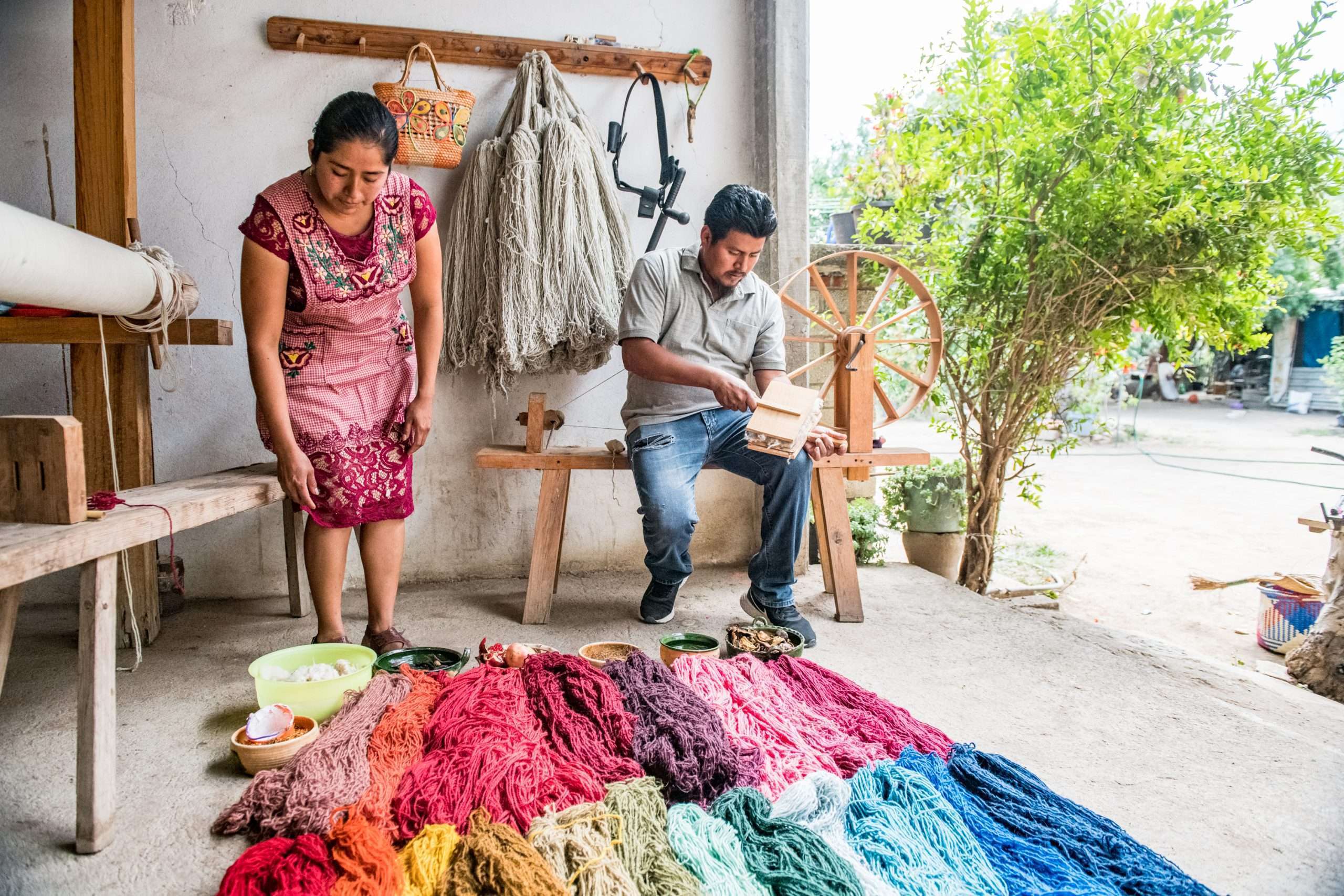 Two Oaxacans stand over a pile of hand-dyed yarns. There are maybe 20 large piles, in vivid reds and blues.