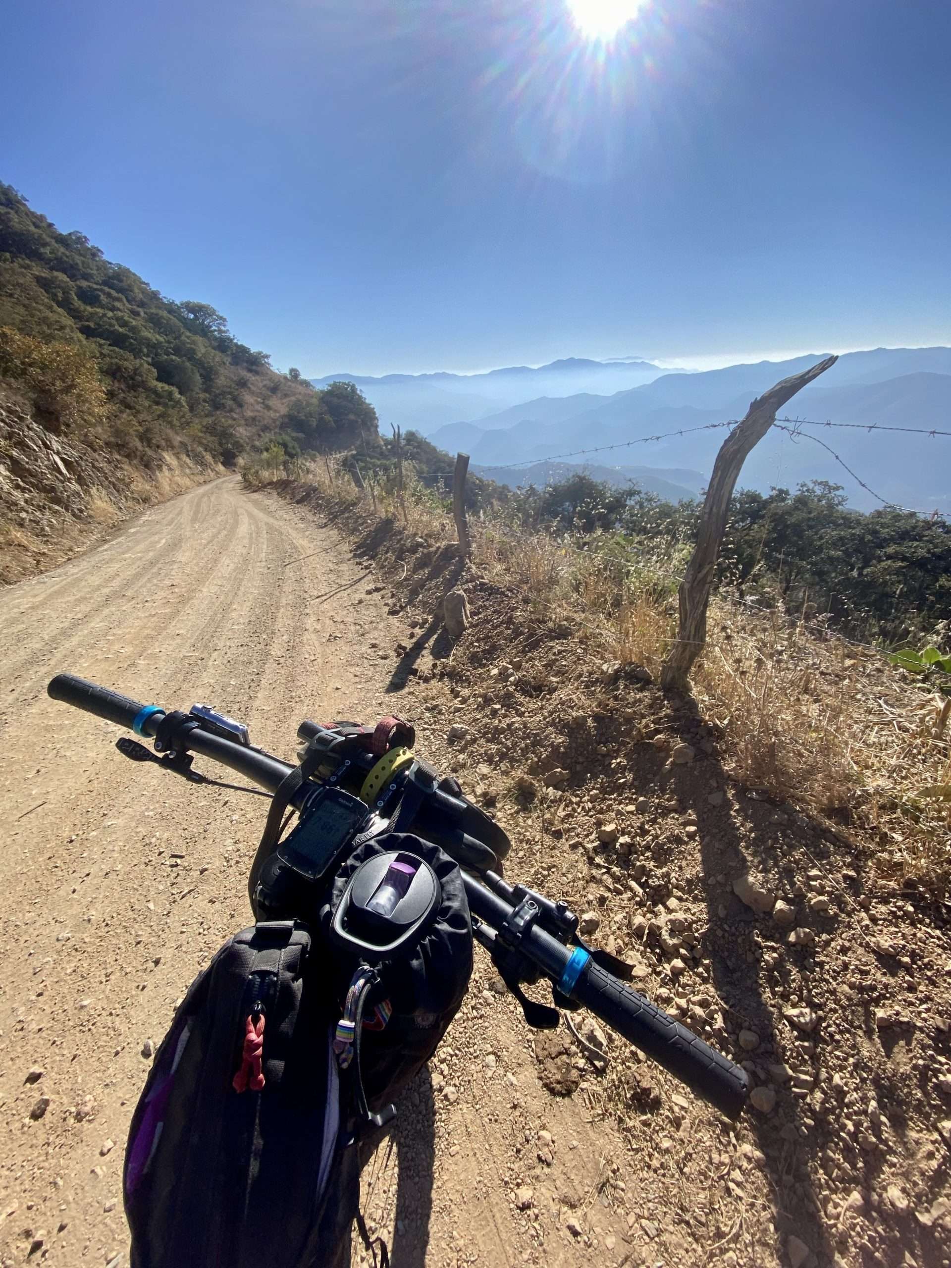 A mountain bike points down a gravel road in a chaparral landscape under a bright blue sky. The sun sparkles at the top of the frame. The road is deserted and the bike has a number of bags attached.