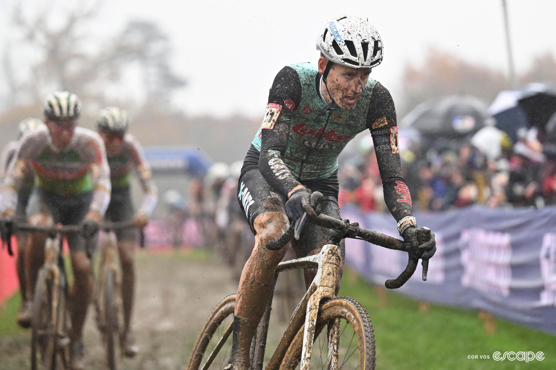 Jens Adams leads a group splattered with mud during CX World Cup Dublin, a large crowd watching the group pass with a number of umbrellas visible.