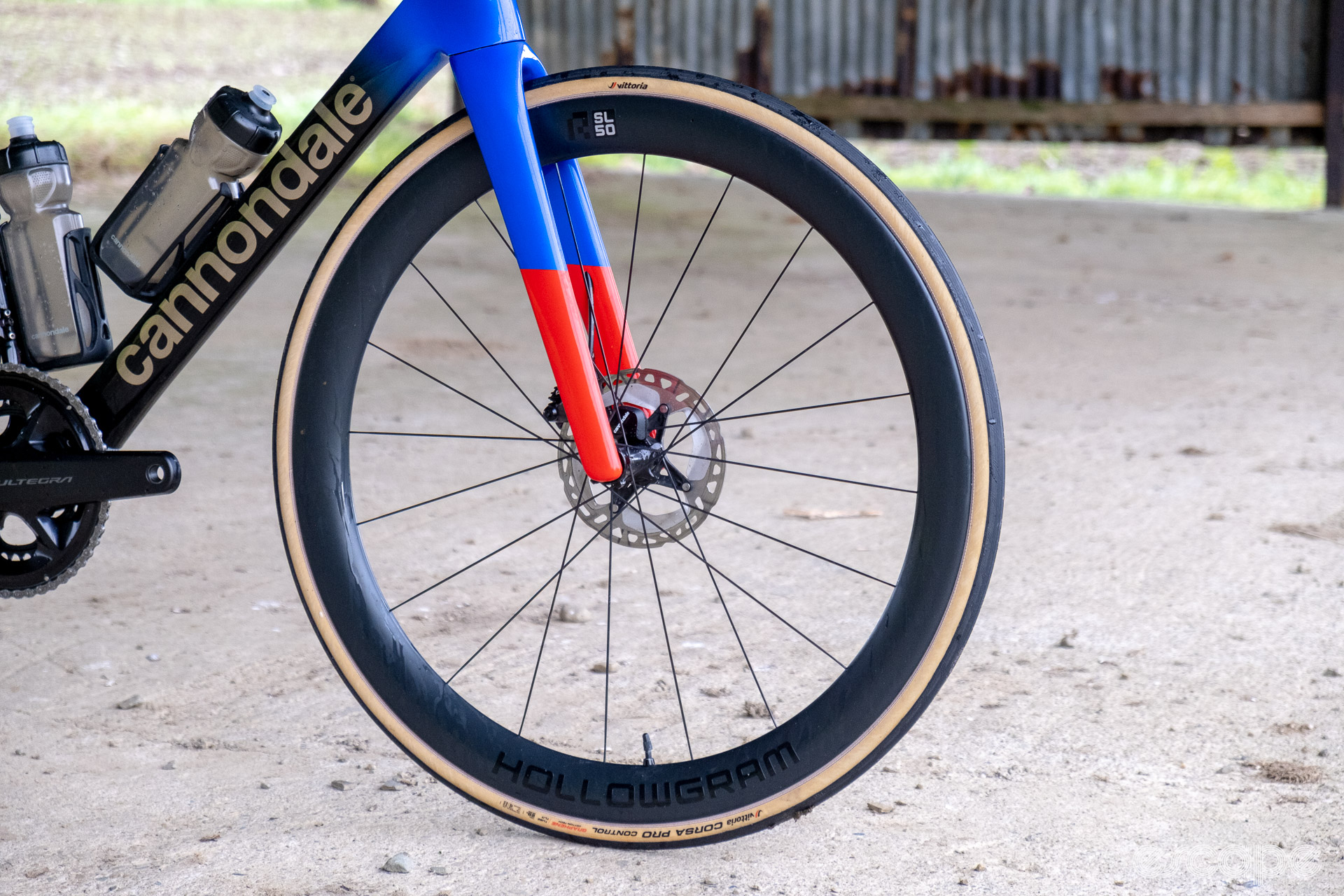 The photo shows the HollowGram and R-SL 50 front wheel on a Cannondale SuperSix Evo Hi-Mod 2 with a Vittoria Corse Pro Control tyre. The picture is taken in an old barn with rusted and bent tin walls and a concrete floor.