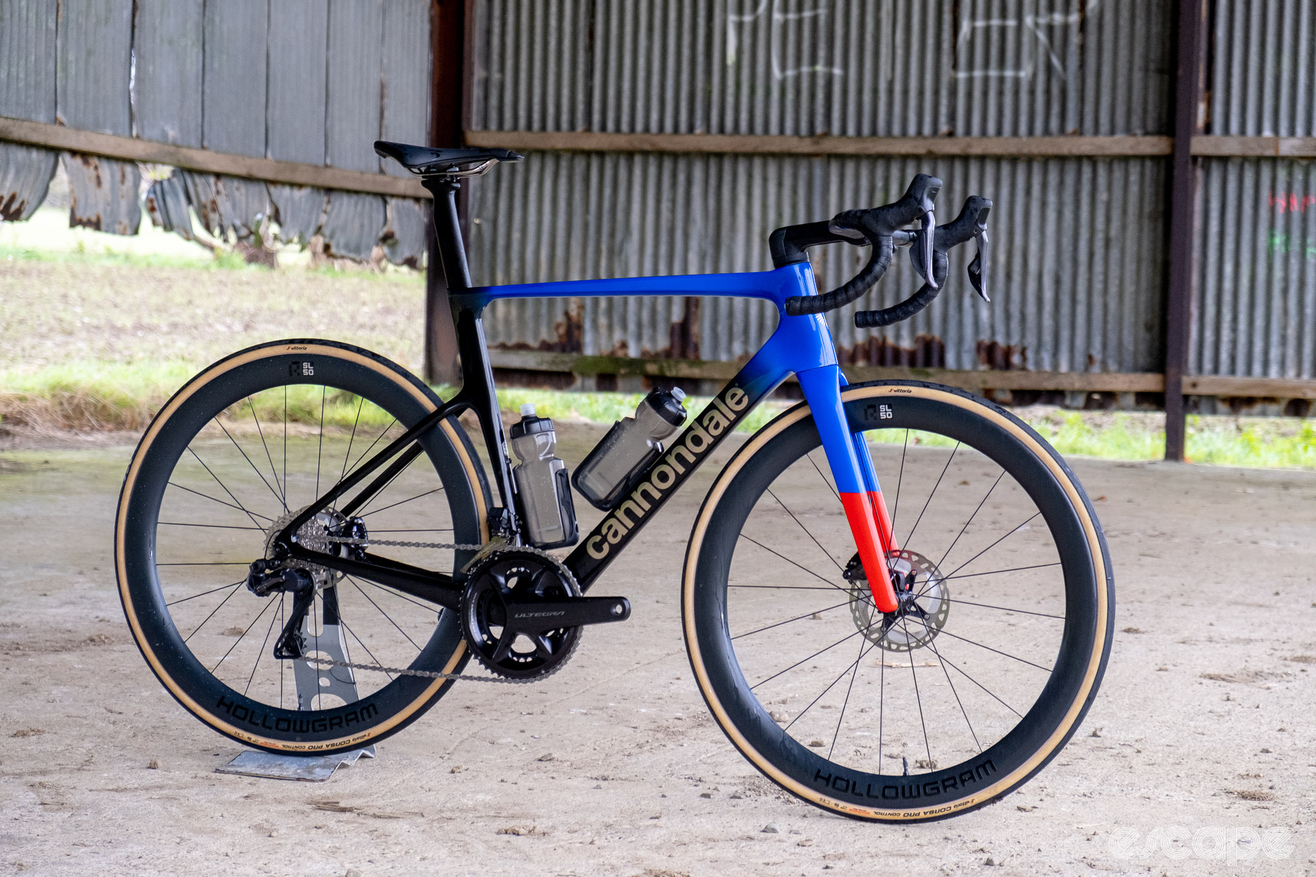 The photo shows a Cannondale SuperSix Evo Hi-Mod 2 frame with Ultegra Di2 HollowGram and R-SL 50 wheelset pictured in an old barn with rusted and bent tin walls and a concrete floor.