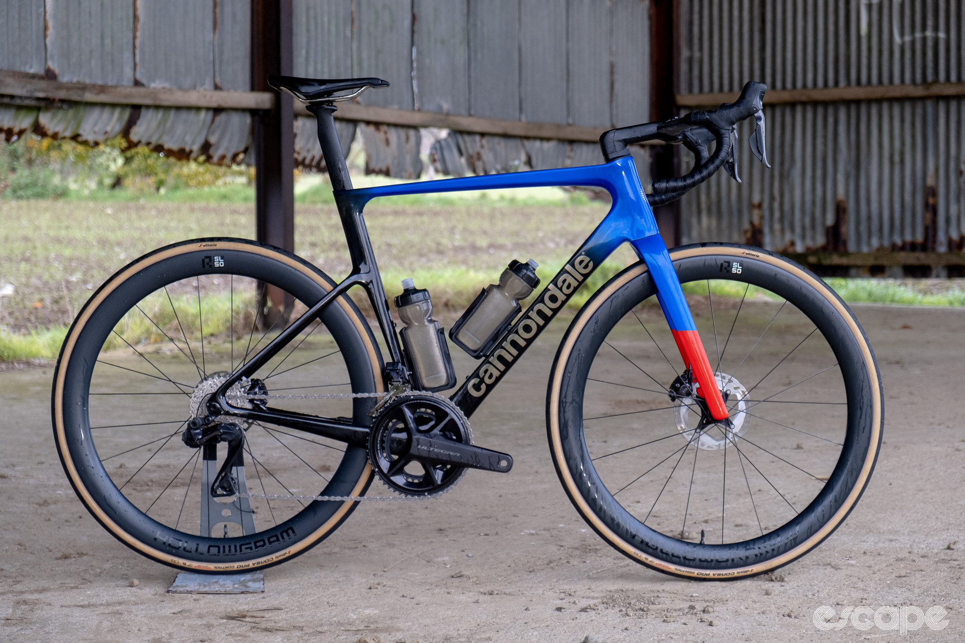 The photo shows a Cannondale SuperSix Evo Hi-Mod 2 frame with Ultegra Di2 HollowGram and R-SL 50 wheelset pictured in an old barn with rusted and bent tin walls and a concrete floor. Rain drops from a torrential shower are running down the front carbon rim.