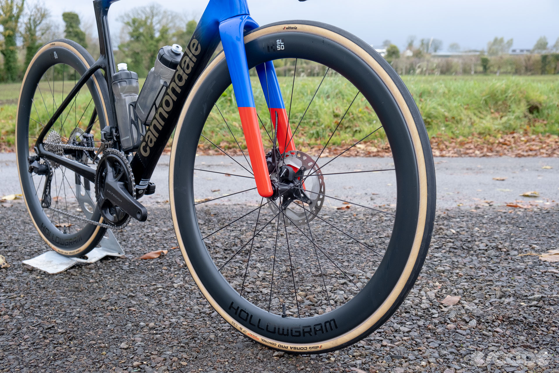 The photo shows the front HollowGram and R-SL 50 wheel and the forks on a new Cannondale SuperSix Evo Hi-Mod 2 along a country lane with a field and trees in the background.