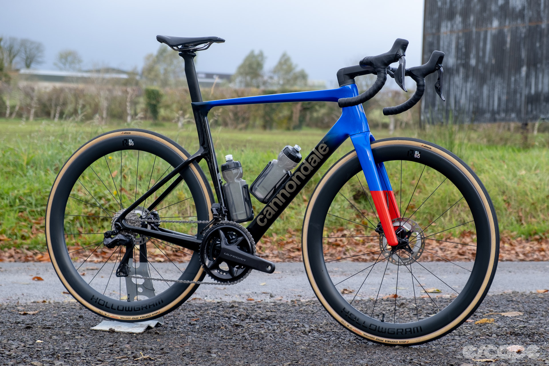 The photo shows a new Cannondale SuperSix Evo Hi-Mod 2 with Ultegra Di2 groupset, HollowGram R-SL 50 wheelset, Prologo saddle, Vision Trimax Carbon Aero handlebars and Conceal stem along a country lane in front of a field and an old barn.