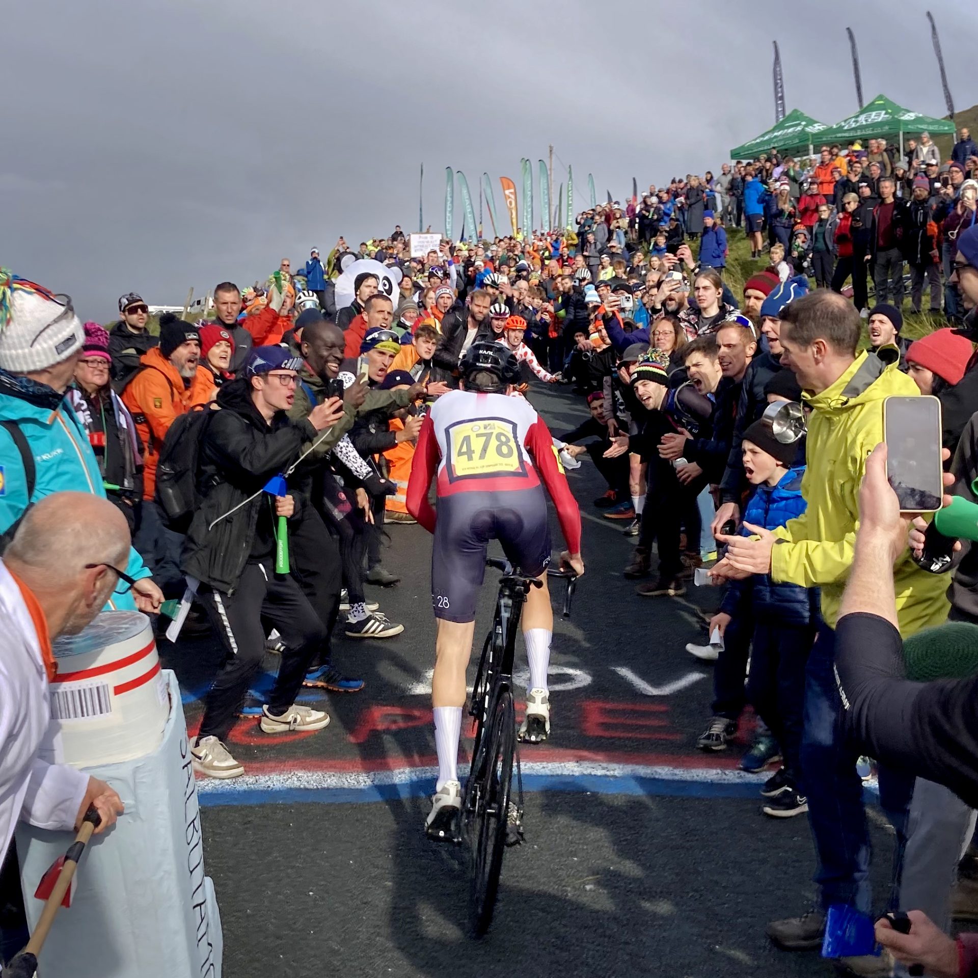 The image shows huge crowds cheering on Ed Laverack as he climbs "The Struggle" road in the 2023 British National Hillclimb championships. The photo shows Laverack from the rear and his race number 478 is clearly visible.