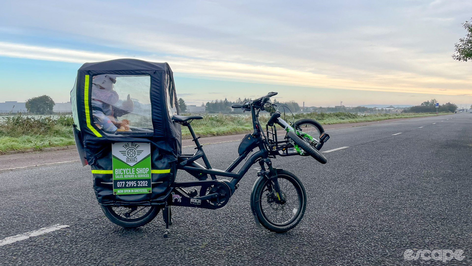 The photo shows a kid sitting on the back of a Tern GSD cargo bike with a kid's bike strapped to the front rack of the GSD.