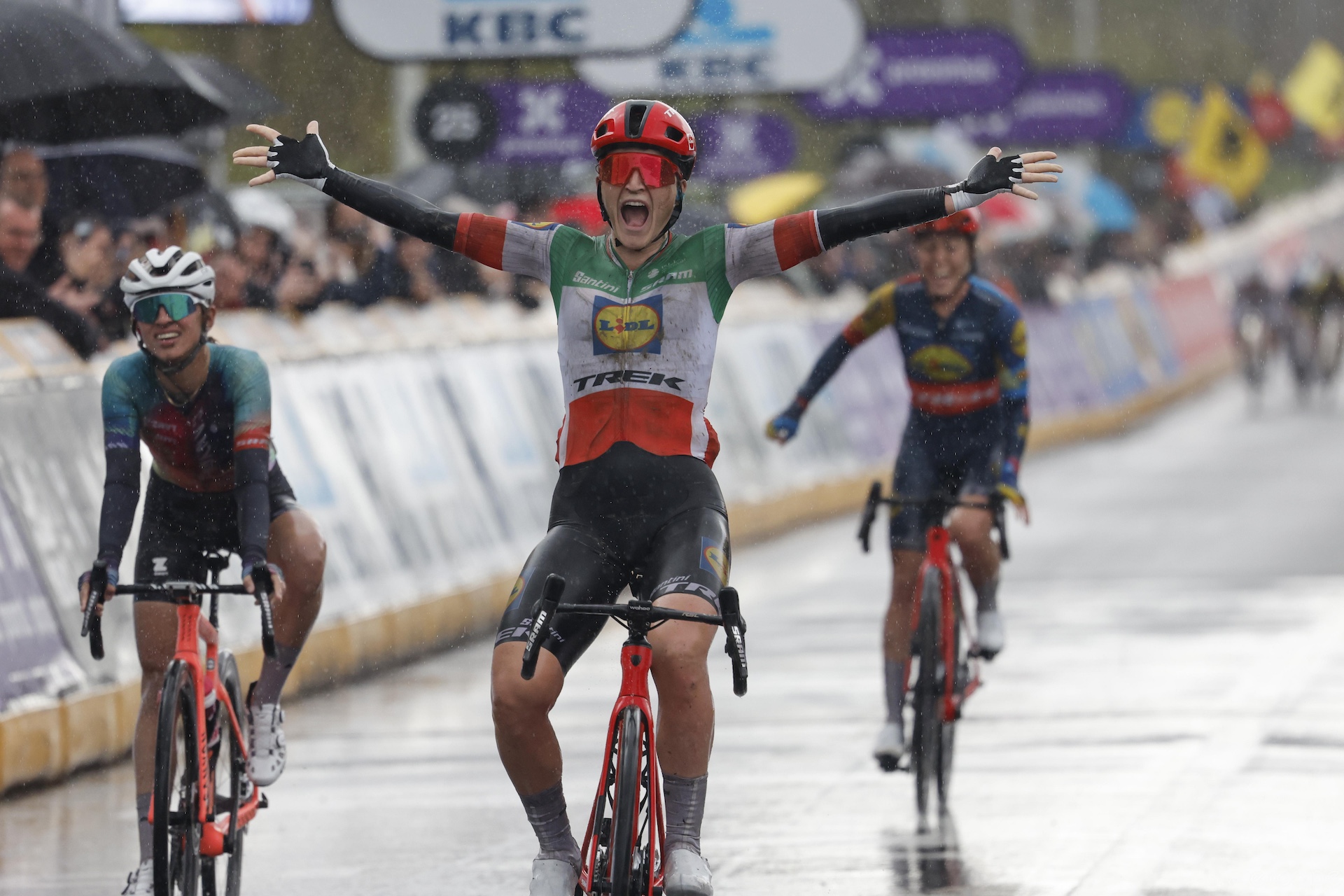 three women covered in mud cross a bike race finish line, the one in the middle raises her arms in celebration