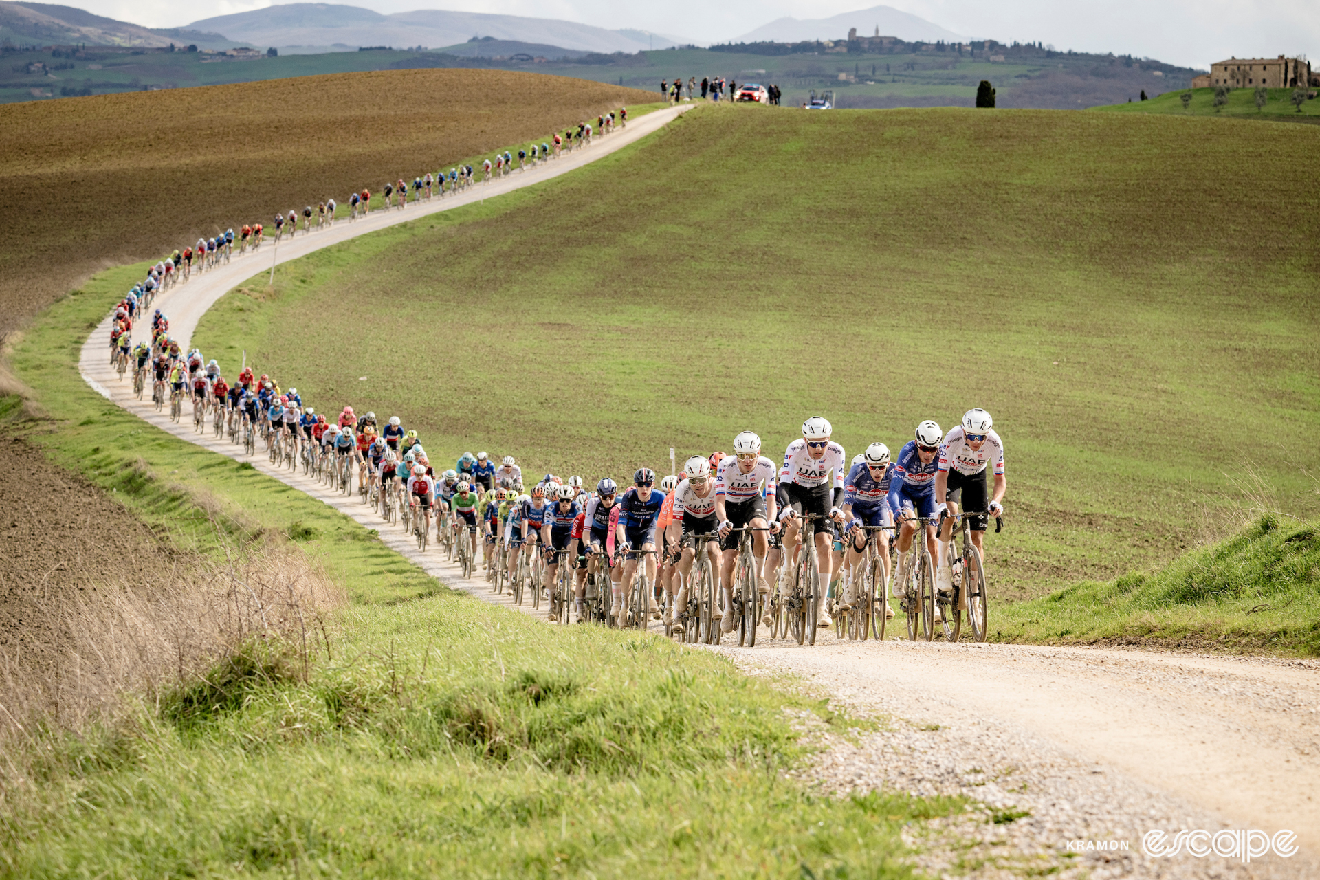 UAE Team Emirates leads the strung-out peloton over the white gravel roads of Strade Bianche against a backdrop of the rolling Tuscan countryside.