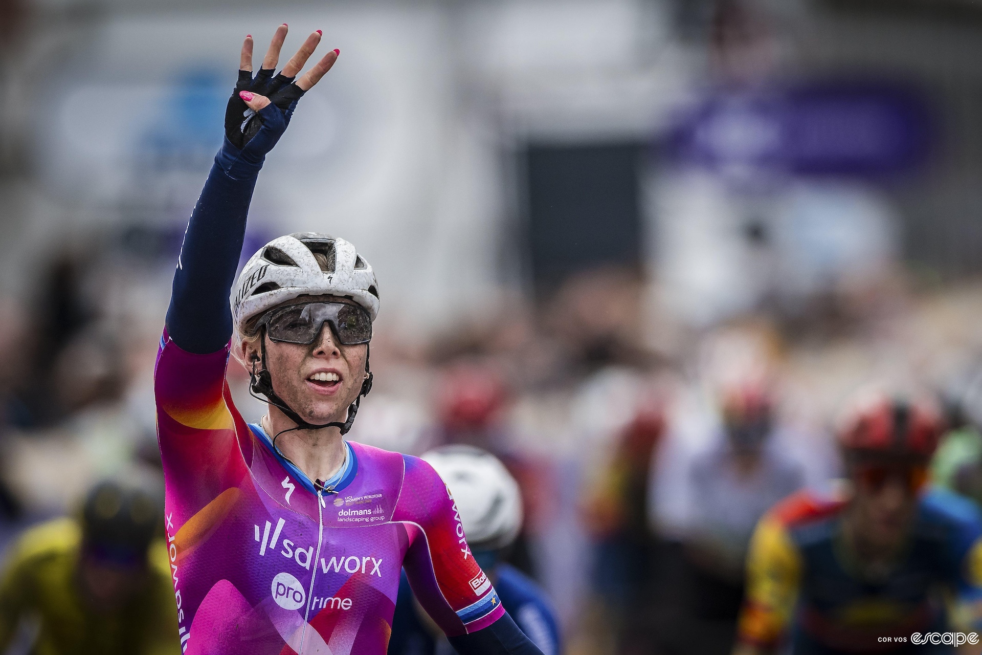 A female cyclist covered in mud raises her arm in victory, holding up four fingers.