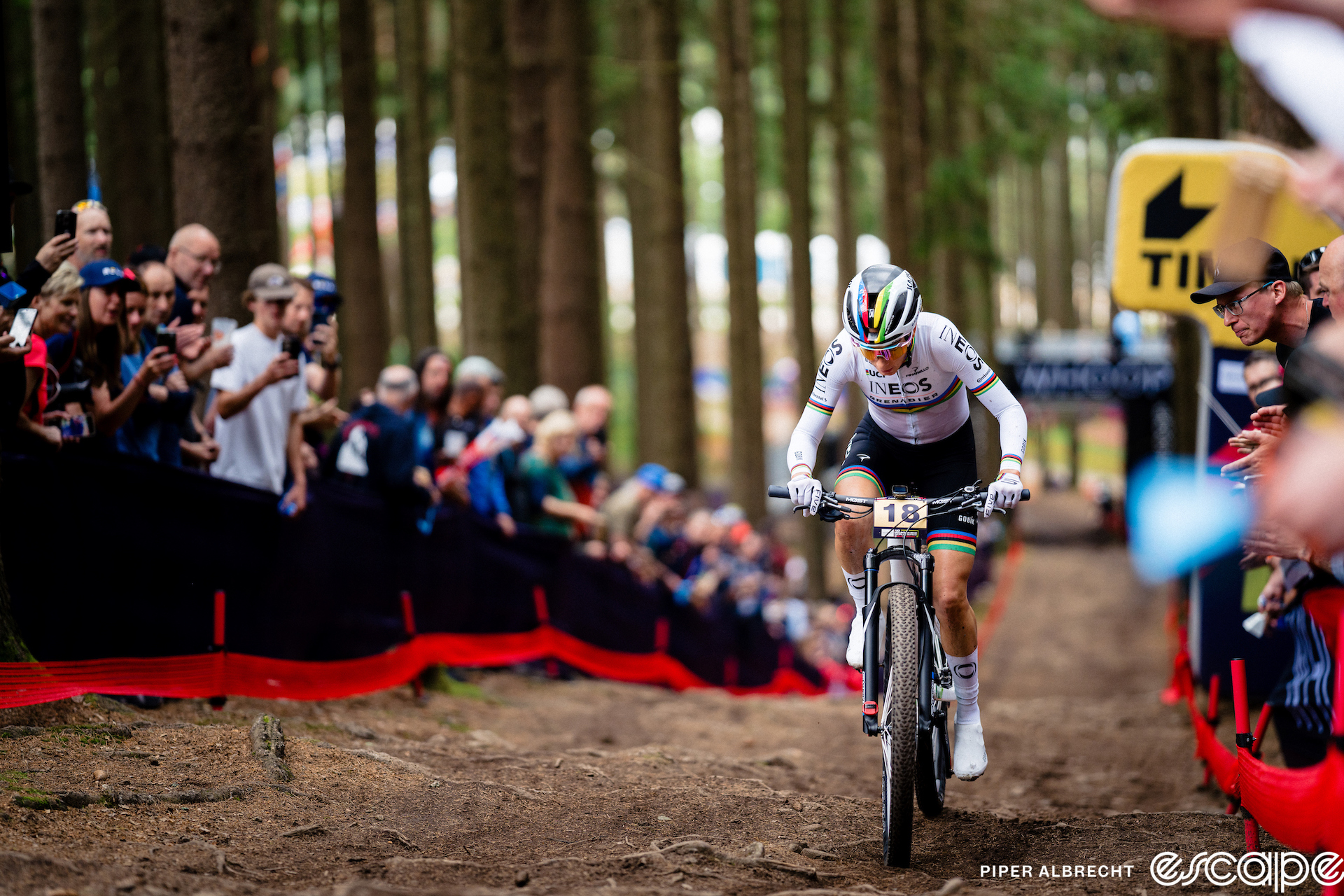 Pauline Ferrand-Prevot climbs a long, straight section at the Nové Mesto World Cup. It's relatively untechnical with some roots, lots of passing lanes, and lined by fans. In the far distance under a sponsor gantry is a lone, blurry rider in pursuit.