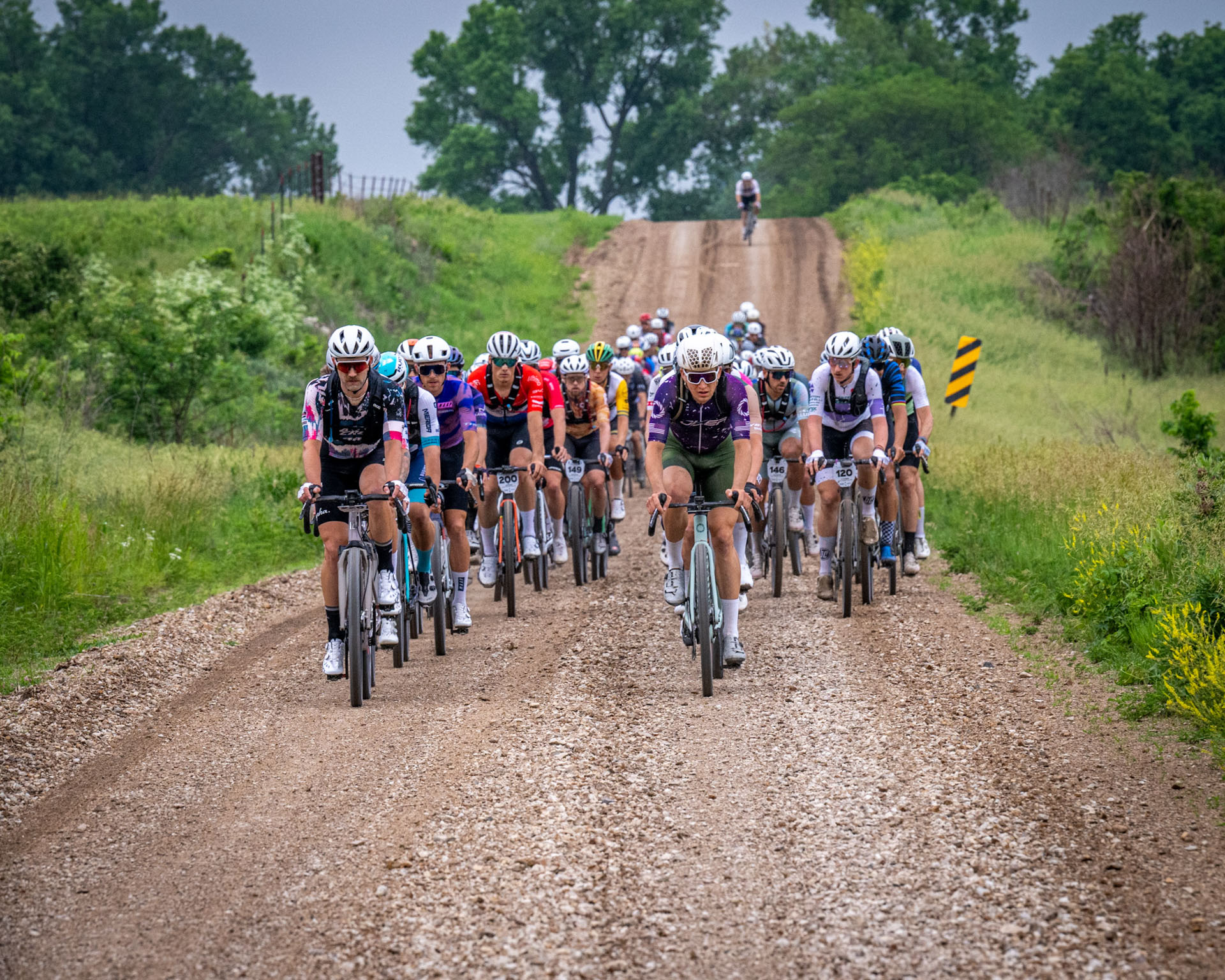 Riders race in a pack at the 2024 Unbound Gravel. They're on a wide dirt road with lush green fields and trees around them.