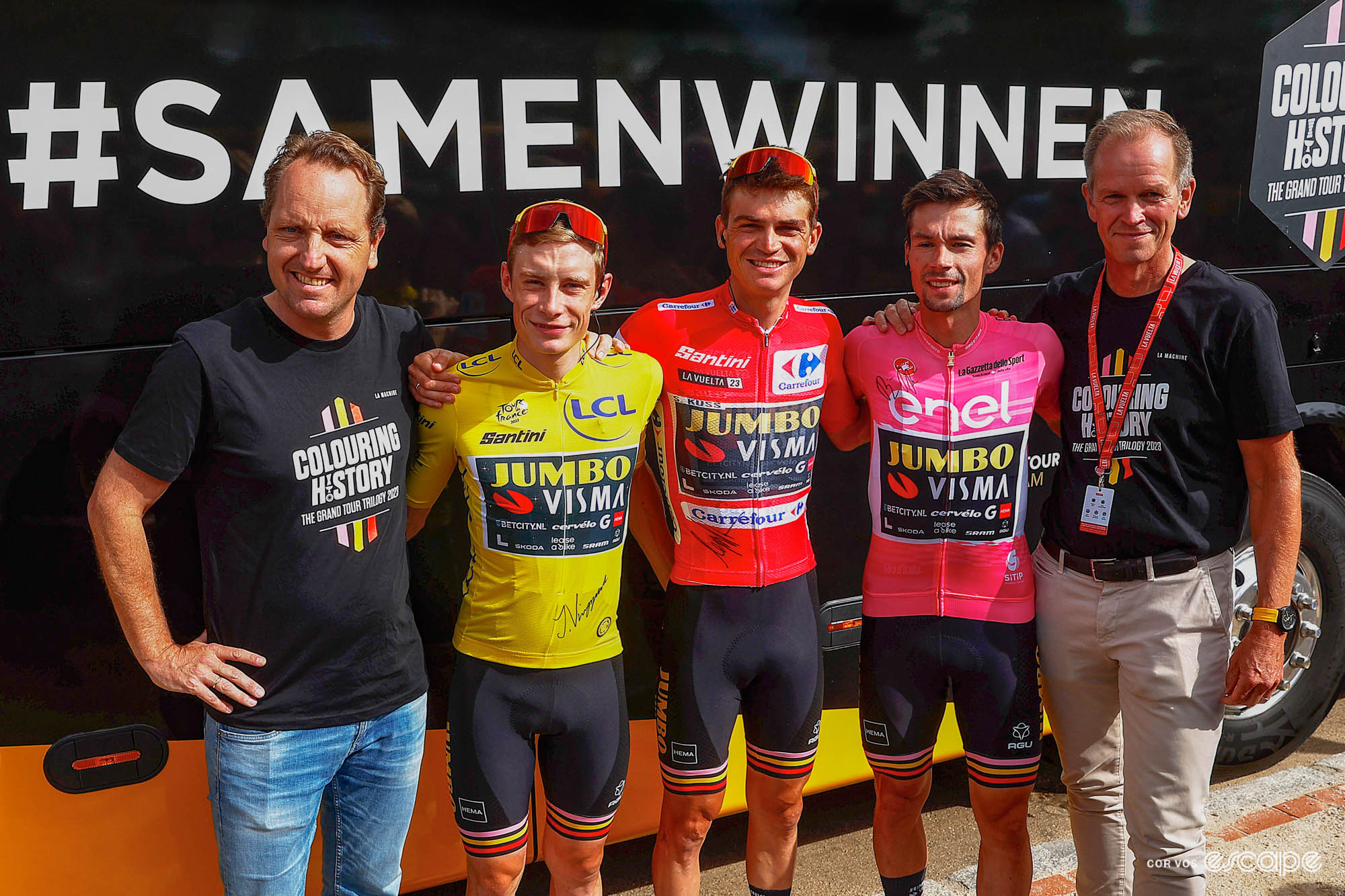 From left to right, Visma director Merijn Zeeman, Jonas Vingegaard, Sepp Kuss, Primož Roglič, and manager Richard Plugge stand for a photo in front of the Visma bus. The three riders are wearing the leaders' jerseys from the Grand Tours. Behind them the phrase #samenwinnen is printed in large letters on the side of the bus.