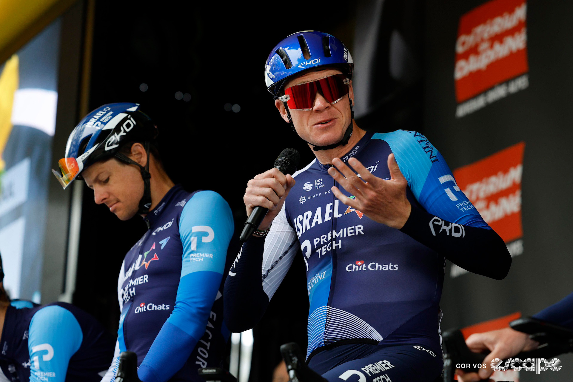 Chris Froome speaks into a microphone on stage before stage 1 of the 2024 Criterium du Dauphiné as teammate Jakob Fuglsang looks down at his stem to Froome's right.