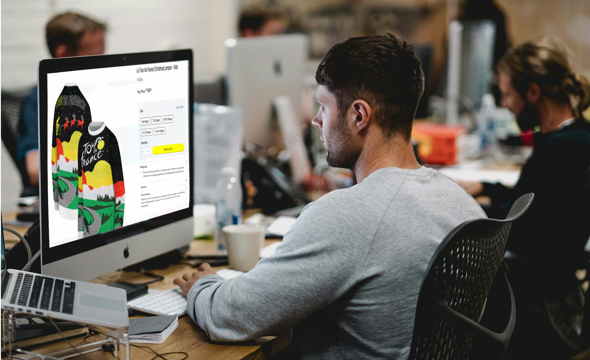 Young male designer sits at his computer in a modern office, with a Tour de France Christmas jumper on his screen.