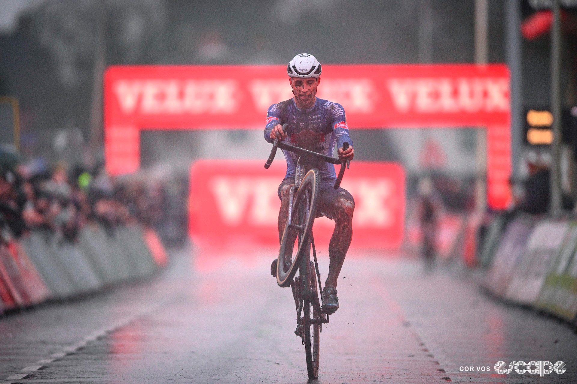 A mud-covered Niels Vandeputte of Alpecin-Deceuninck pops a wheelie as he crosses the line victorious at World Cup Hulst in pouring rain.