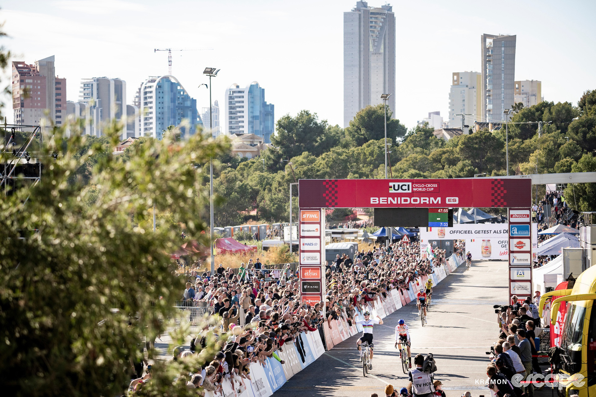 A wide shot of the finish line and the large crowd as Fem van Empel celebrates a narrow victory over Lucinda Brand at cyclocross World Cup Benidorm, the city's towers in the background.