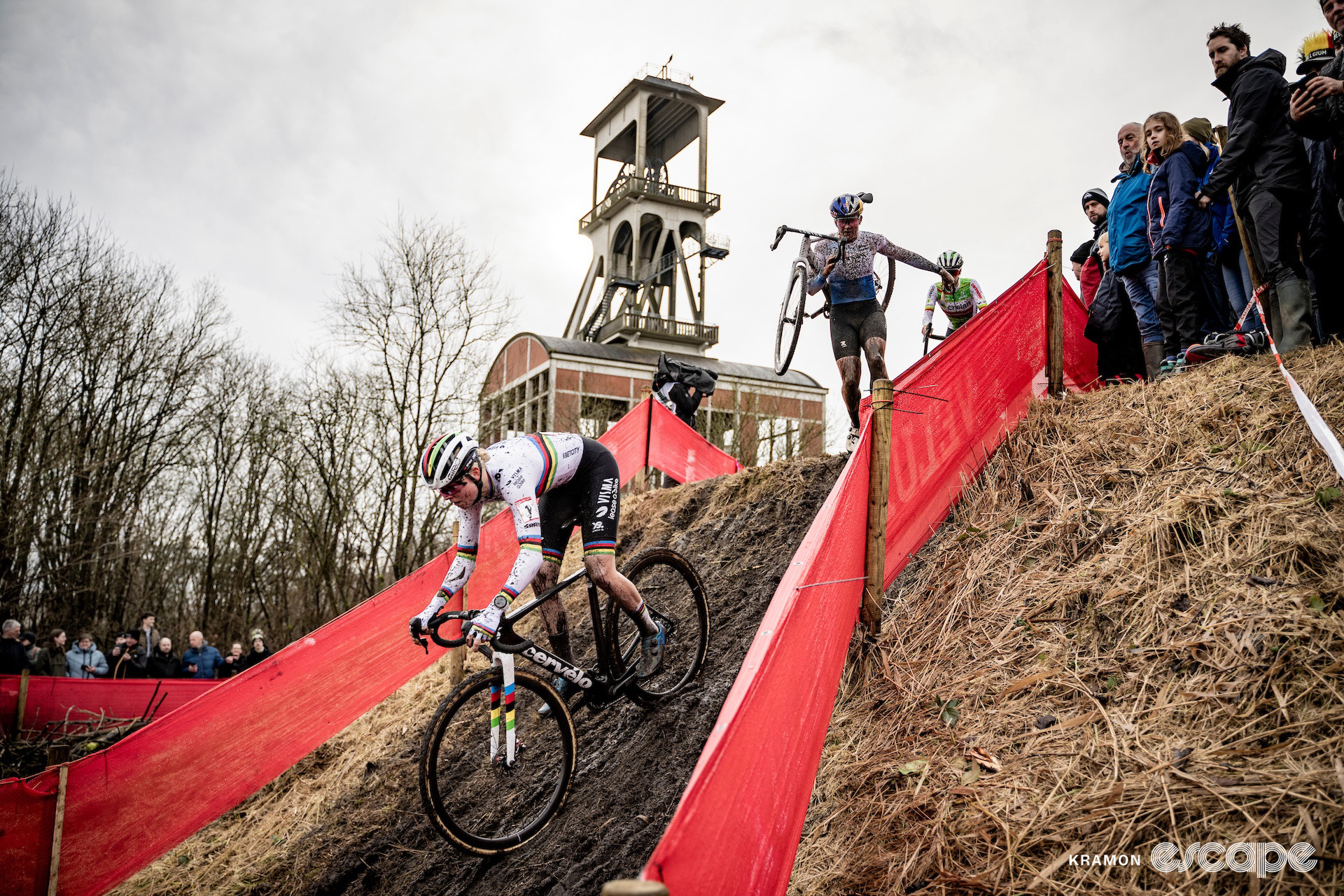 World champion Fem van Empel leads a small group during cyclocross World Cup Maasmechelen, Zoe Backstedt chasing with her bike on her shoulder, distinctive old mining infrastructure towering in the background.