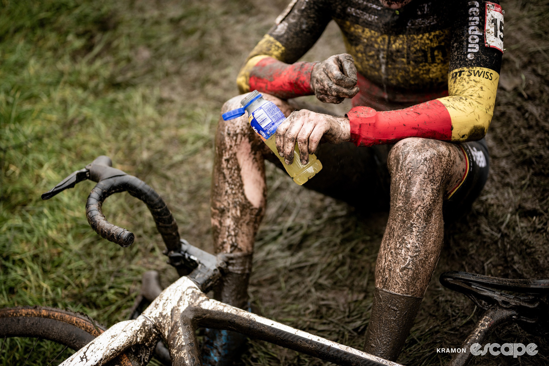 Belgian national champion Marion Norbert Riberolle, pictured from shoulders down, sitting on the muddy grass with her bike after the finish as she rinses mud from her legs with an orange recovery drink.