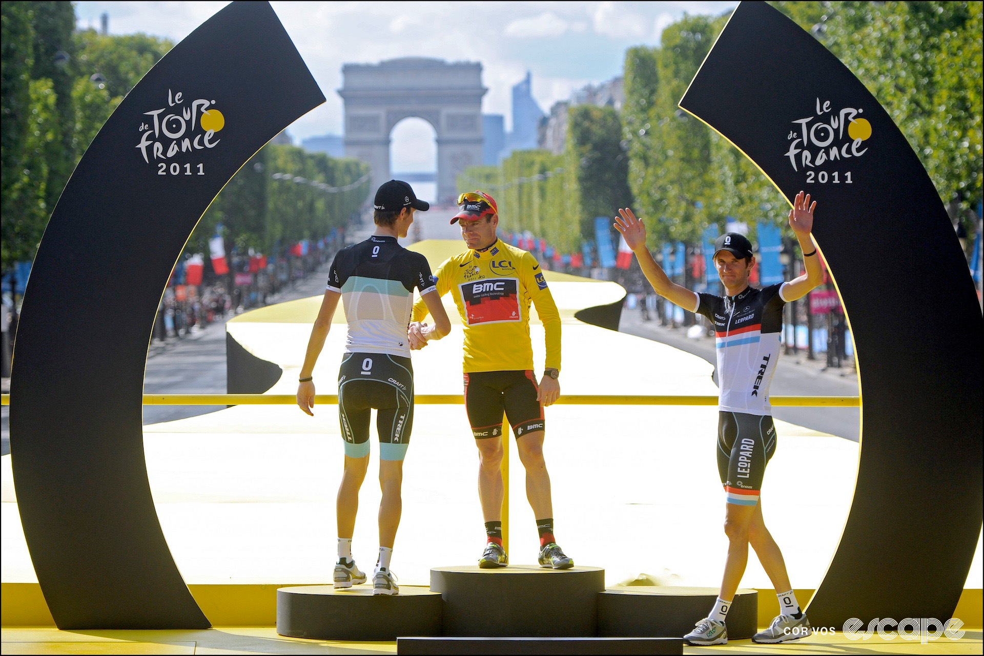 Cadel Evans in the yellow jersey, shaking the hand of Andy Shleck as Frank Schleck waves to the crowd on the final GC podium of the 2011 Tour de France in Paris.