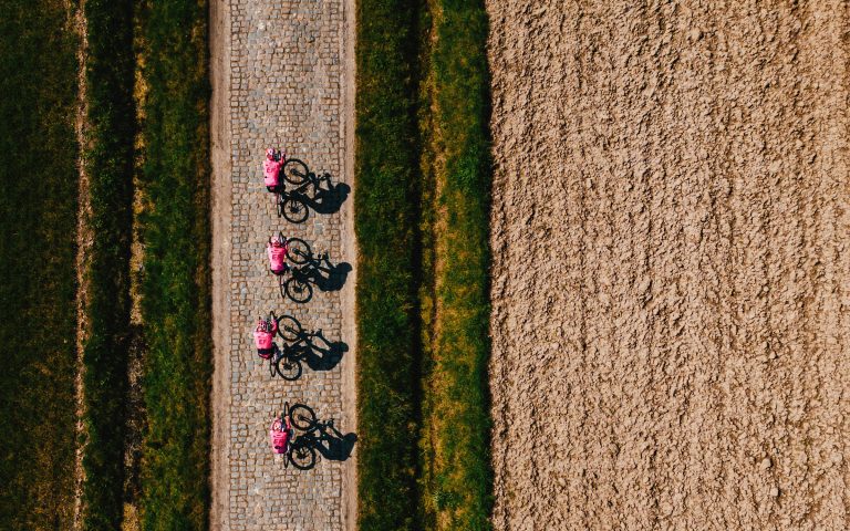 An overhead drone shot of the EF Education-Easypost team on a Paris-Roubaix recon ride. Four riders in distinctive pink EF kits ride in a paceline on a narrow cobbled road next to a freshly plowed field.