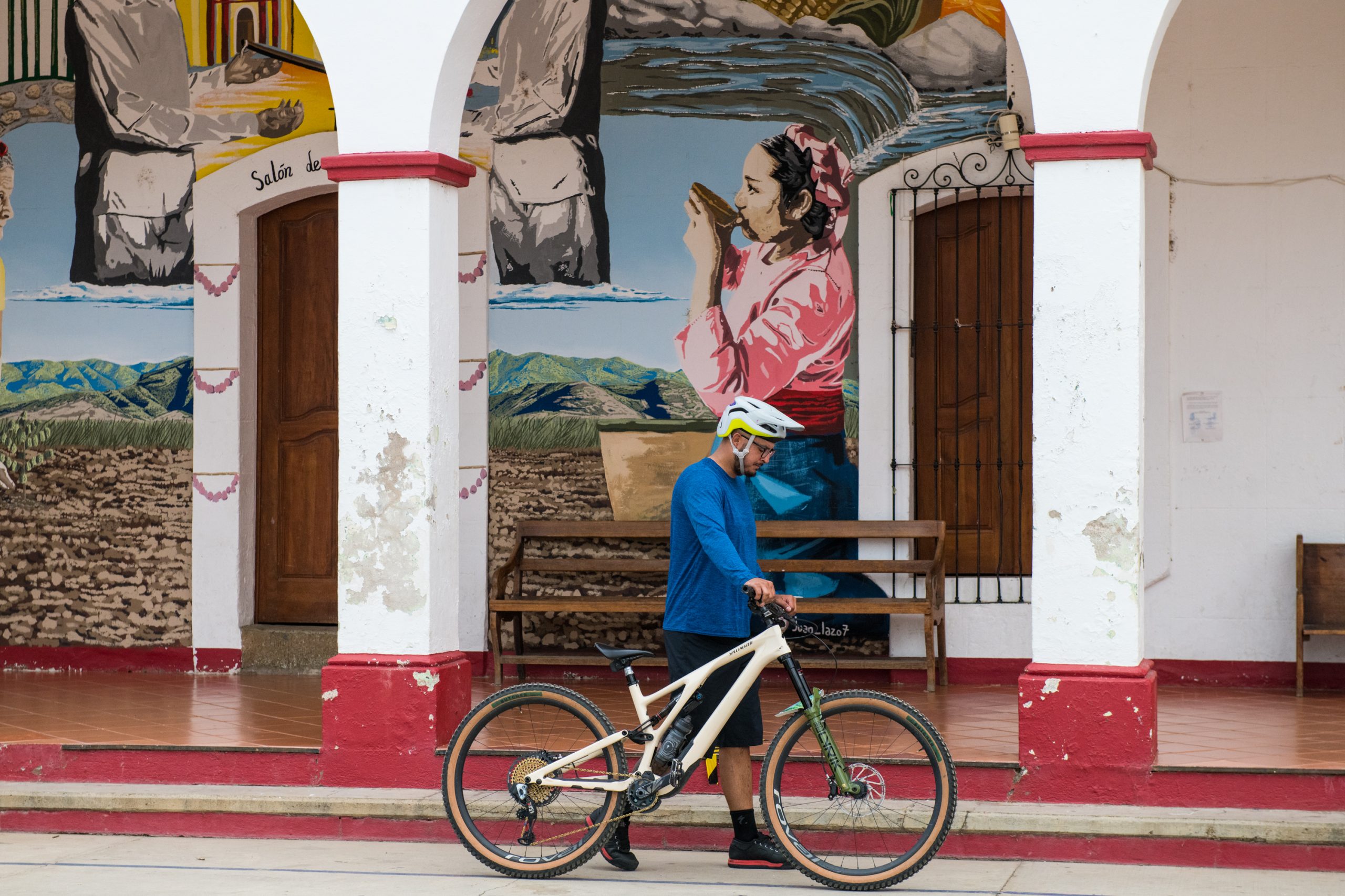 A mountain biker walks his bike in front of a mural of a local woman in a bright pink blouse drinking from a bowl, set against a pastoral scene of farm fields and low hills behind.