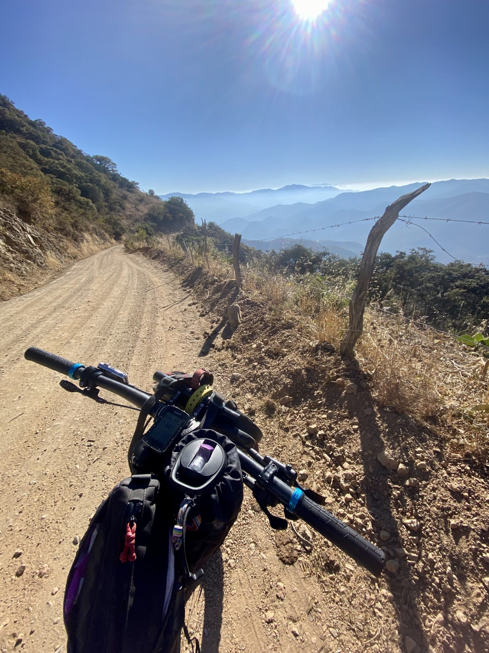 A mountain bike points down a gravel road in a chaparral landscape under a bright blue sky. The sun sparkles at the top of the frame. The road is deserted and the bike has a number of bags attached.
