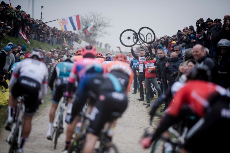 Riders on a cobblestone sector at Paris-Roubaix, passing a line of spectators and team helpers with wheels held aloft.