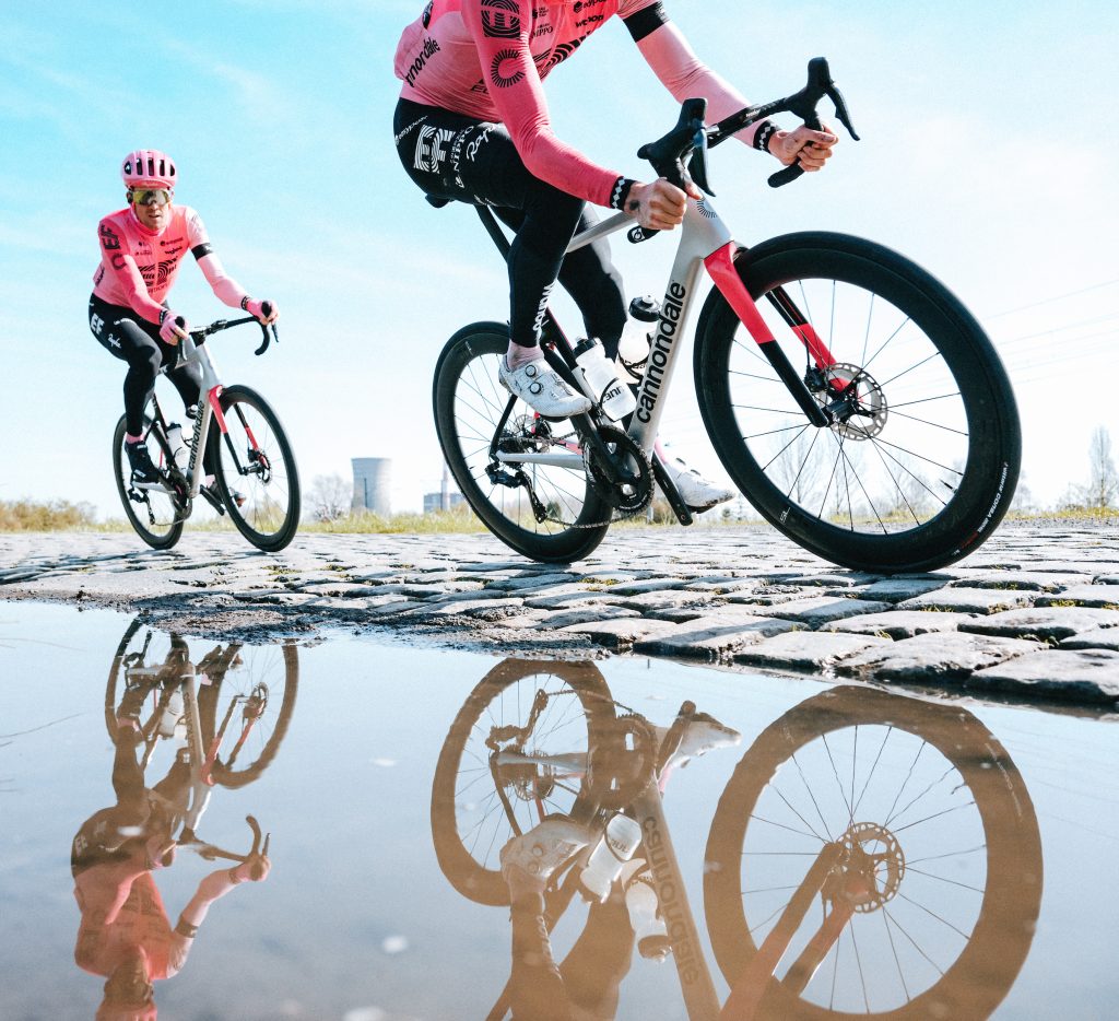 Two EF riders navigate cobbles past a still puddle of water. Their bikes are reflected in its surface. Behind them in the distance is a cooling tower for a nuclear power plant.