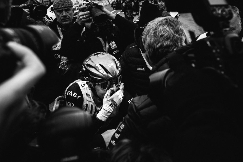 A black and white photo of 2023 Tour of Flanders winner Tadej Pogačar, surrounded by media at the finish. He has a hand over his mouth, as if overcome with emotion at the accomplishment.