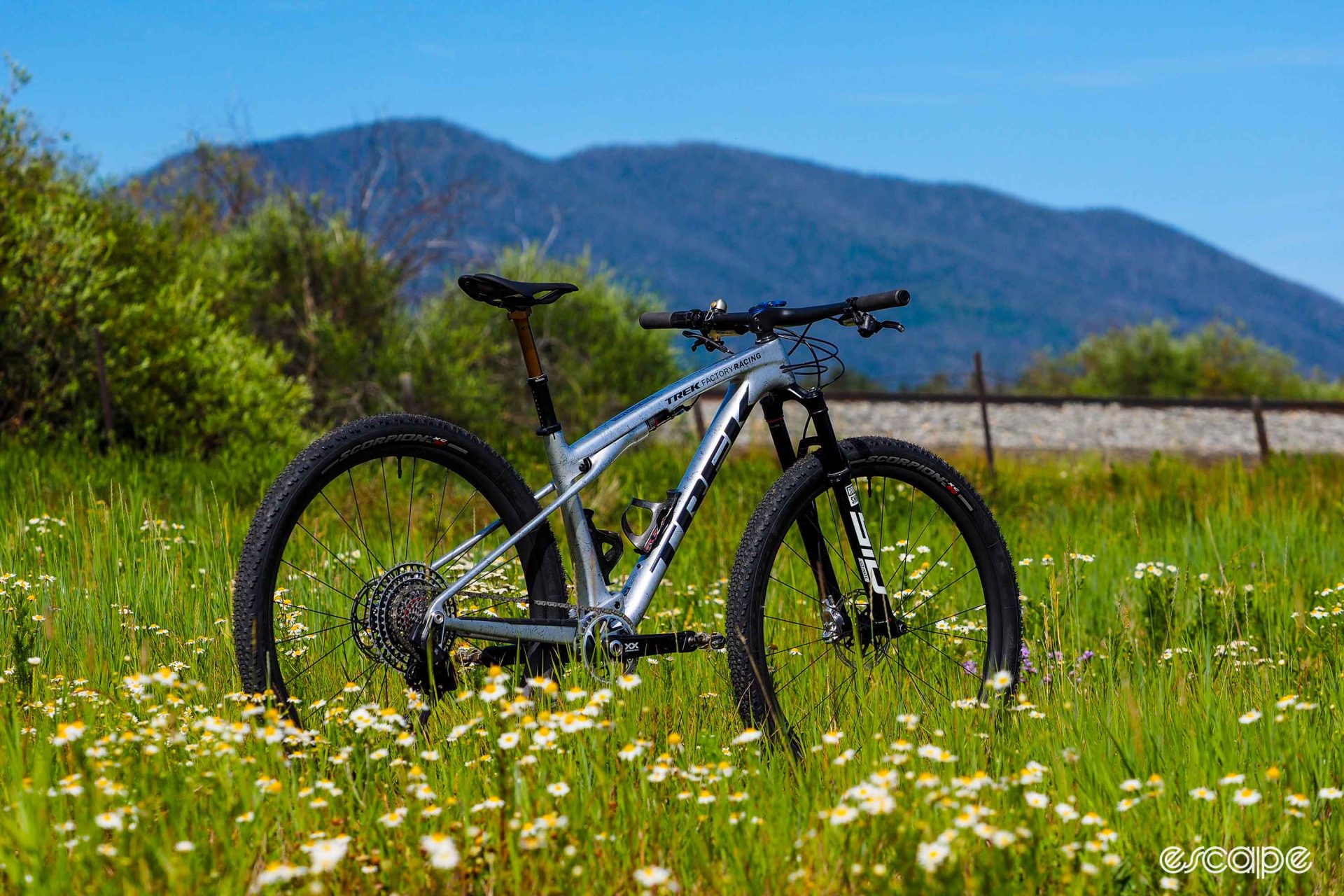 A metallic gray Supercaliber with black logos appears in a field of wildflowers, with a railroad grade and a green forested hill in the background.
