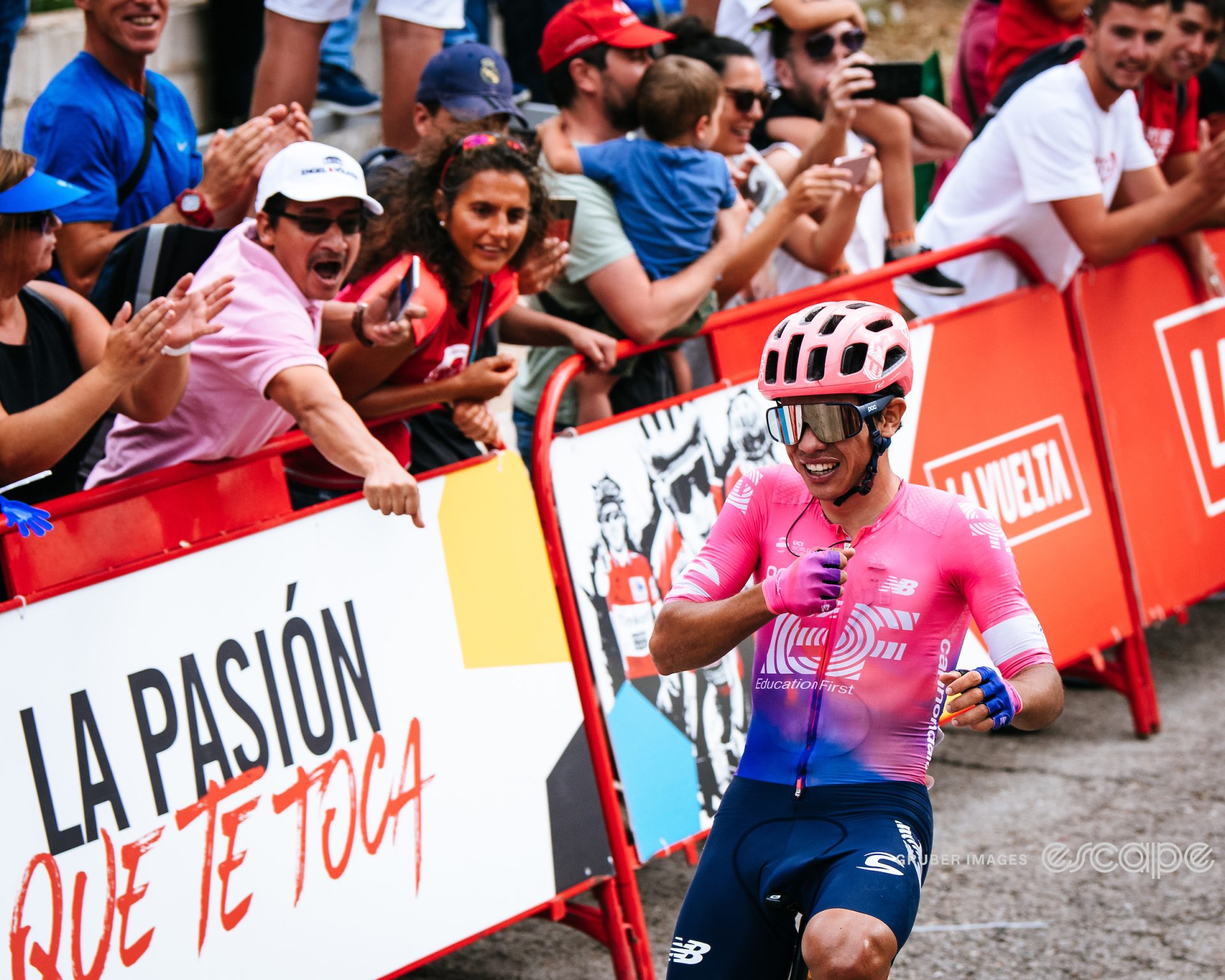 Sergio Higuita crosses the finish line to win stage 18 of the 2019 Vuelta a España. Higuita is wearing the distinctive pink jersey of his EF Education team and is pounding his chest as he crosses the line. Behind, fans lean over the barriers to cheer at him.