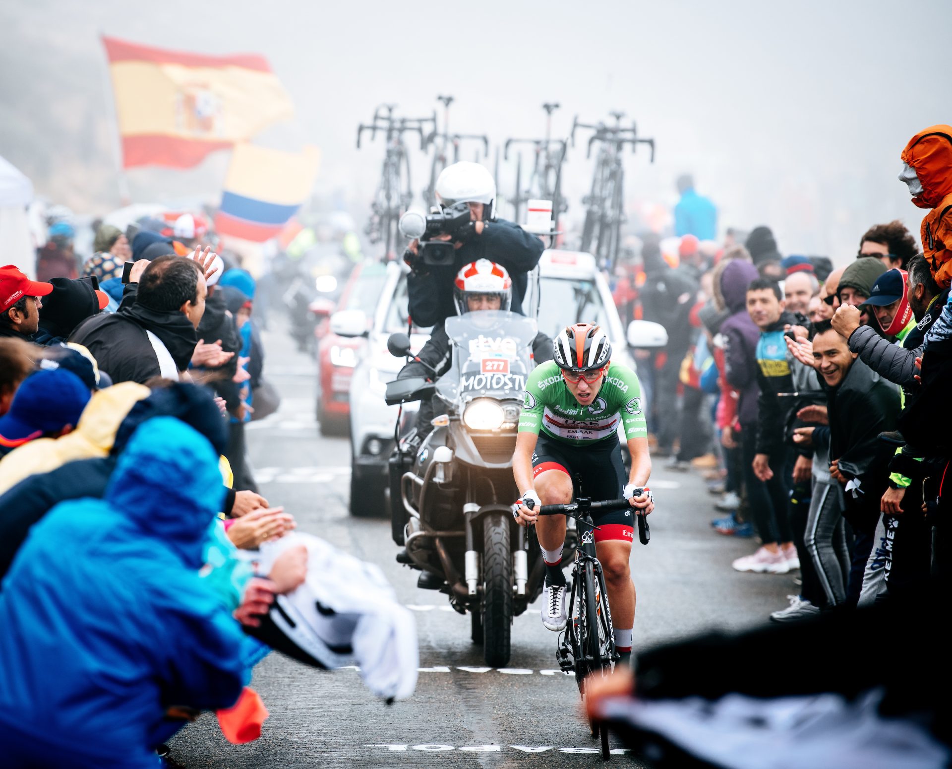 Tadej Pogačar races to the win on stage 20 of the 2019 Vuelta a España on Plataforma de Gredos. He's alone, dressed in the green jersey of the race's best young rider. Fans crowd in on all sides and a TV moto and team car trail him. The road is shrouded in mist and rain, and the fans are dressed in rain gear as flags whip straight out in the wind.