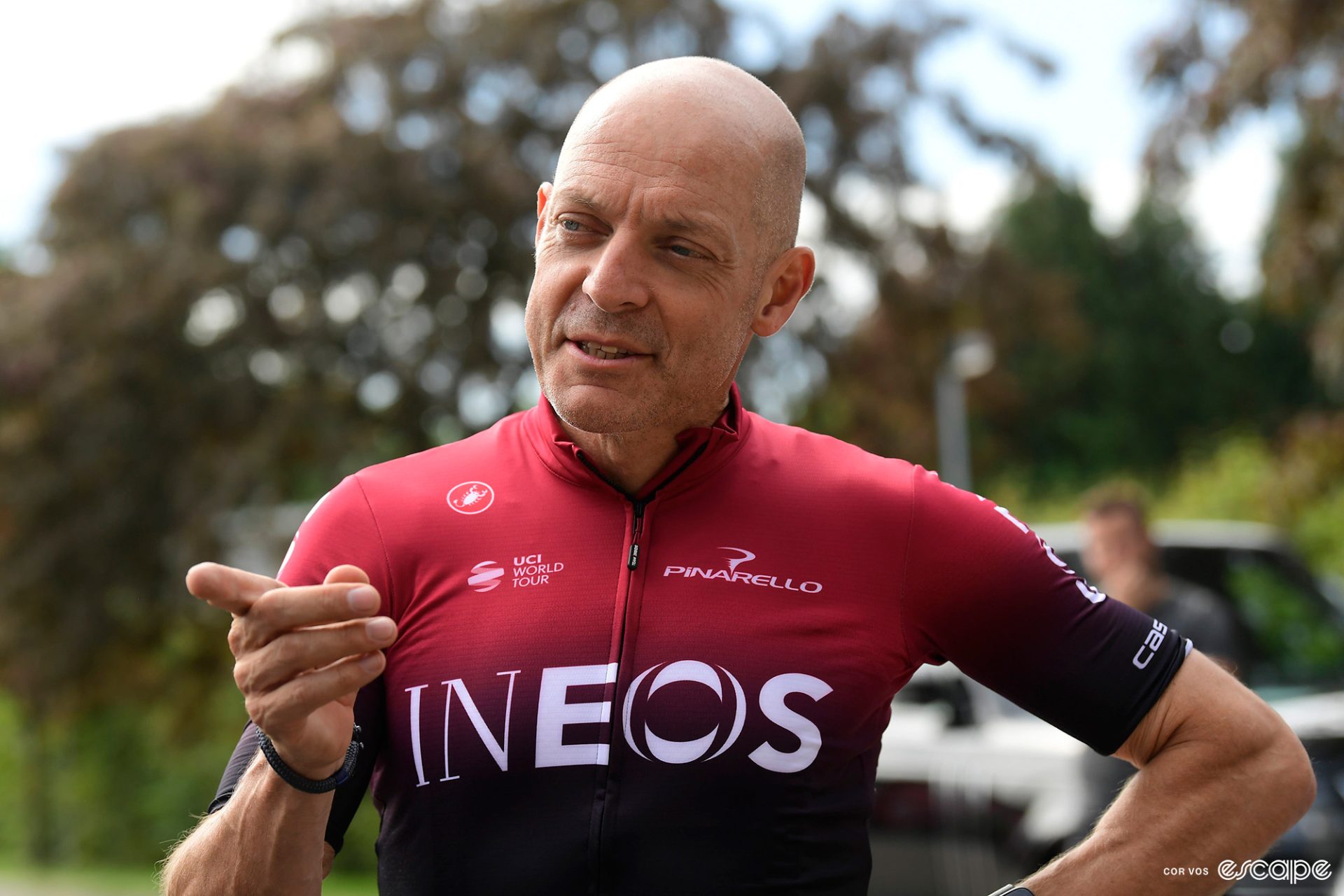 Ineos Grenadiers' Director of Sport, Dave Brailsford, is dressed in a red and black fade Ineos cycling kit. He's pointing to something off-camera and has an especially smarmy look on his face.