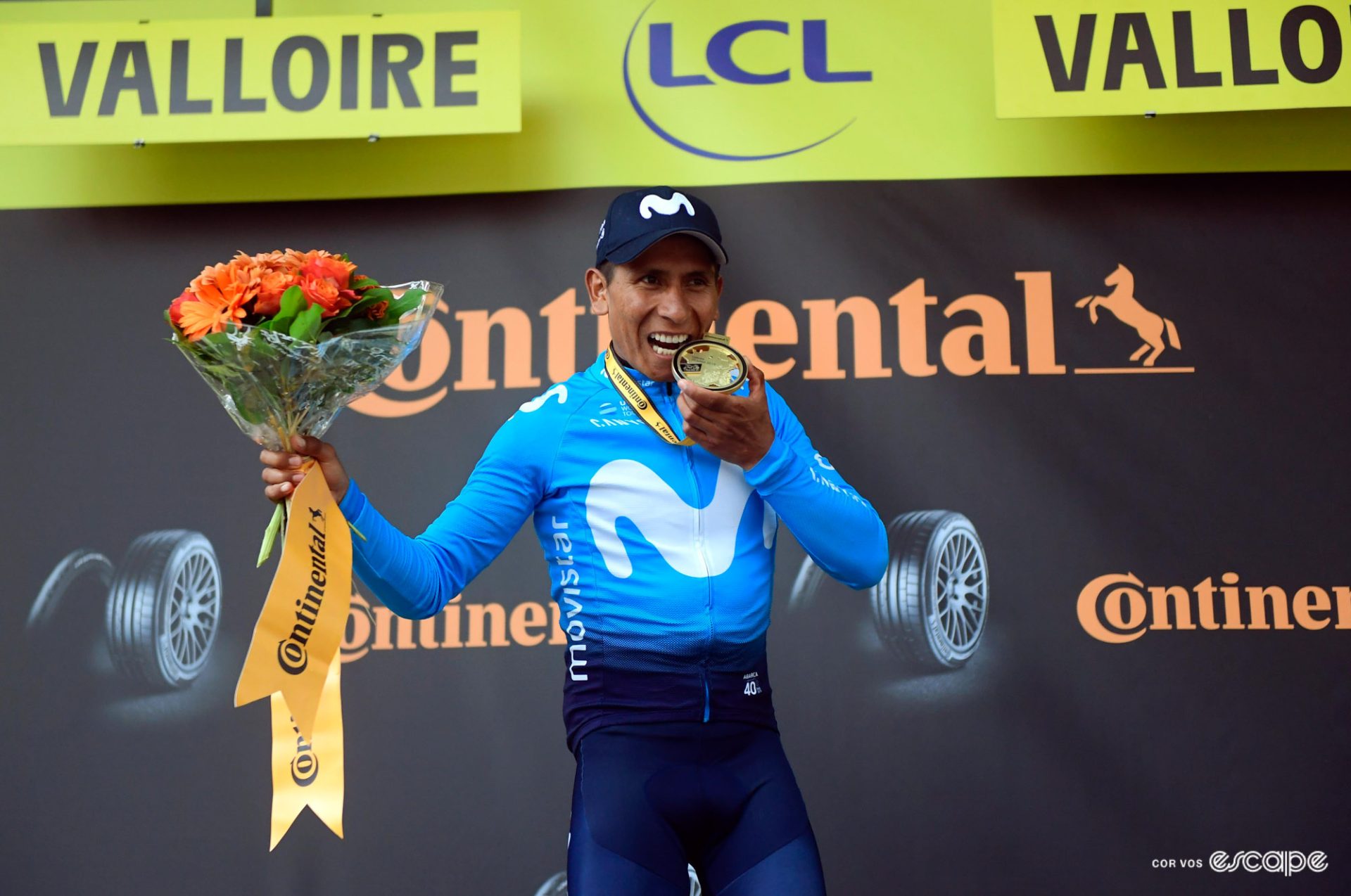 Nairo Quintana on the podium at the 2019 Tour de France having won the stage into Valloire. He is biting a winner's medal and smiling. 