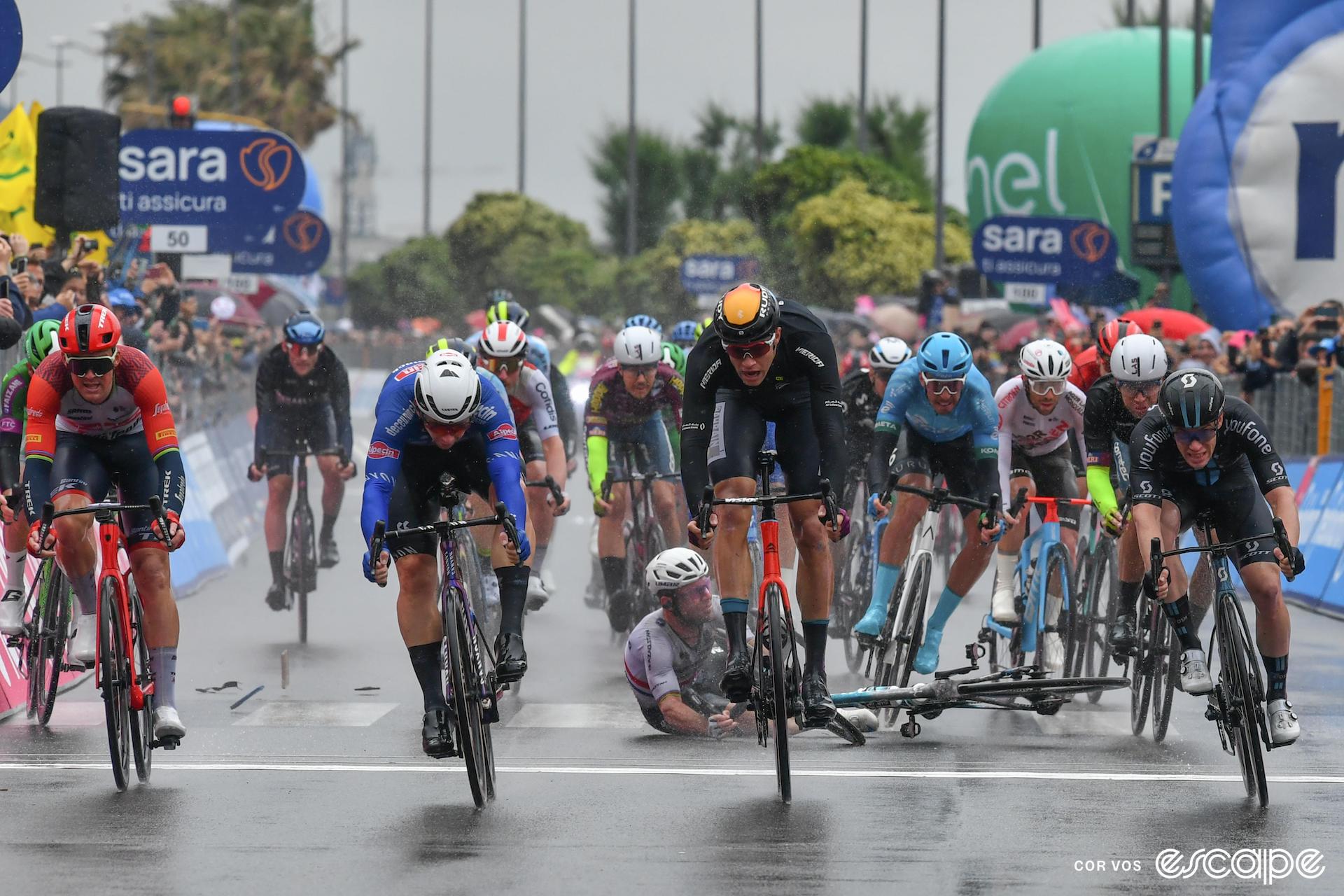 A bunch of pro cyclists sprint for the finish line on a wet road. One of the riders, Mark Cavendish, is hitting ground at the moment the image was taken, his bike lying beside him on the ground.