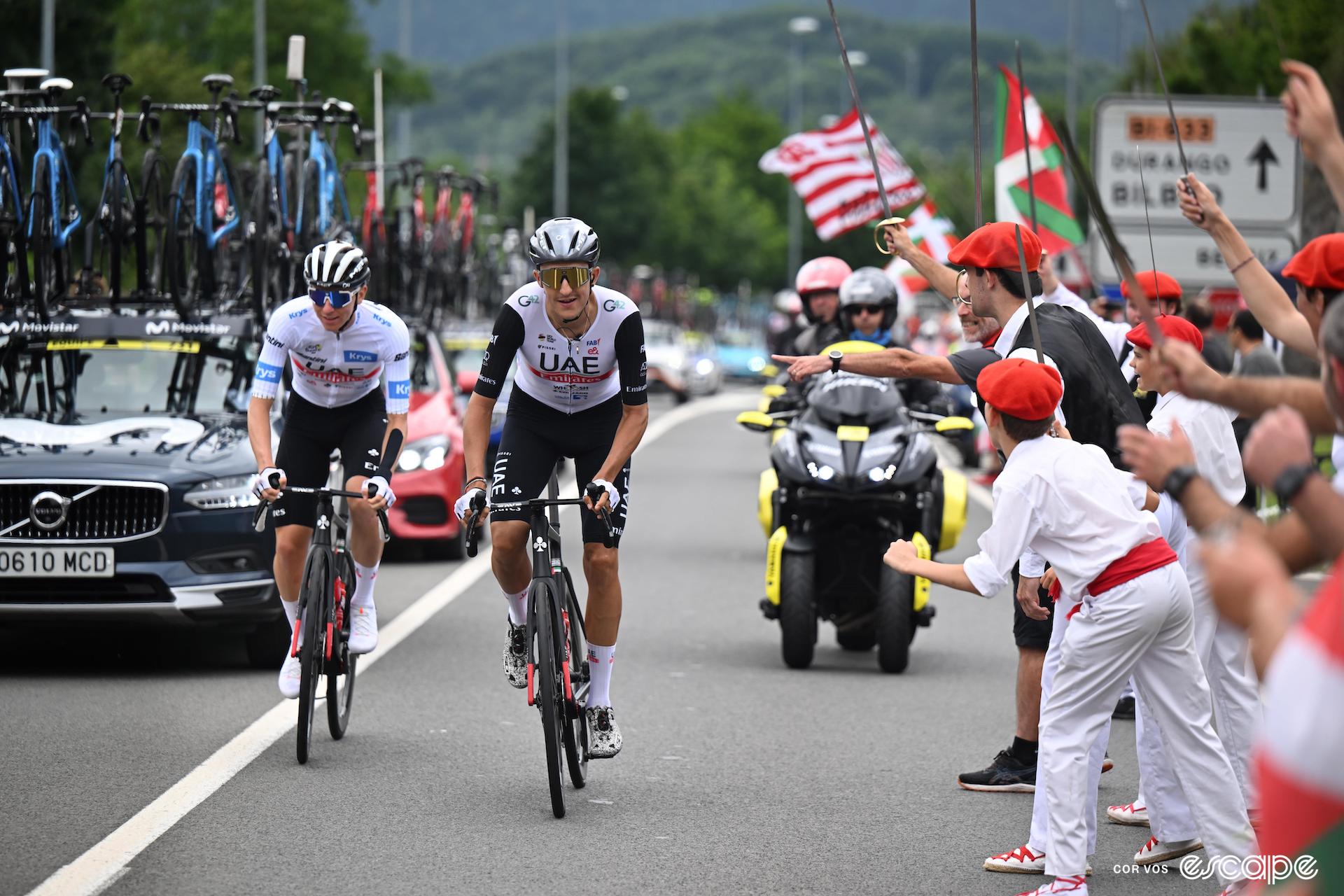 Dane Mikkel Bjerg rides in front and slightly to the right of his teammate, Tadej Pogačar, in an early stage at the Tour de France. Pogačar is in the white jersey of best young rider. The roadside is packed with Basque fans, including a number wearing the traditional txapela beret.