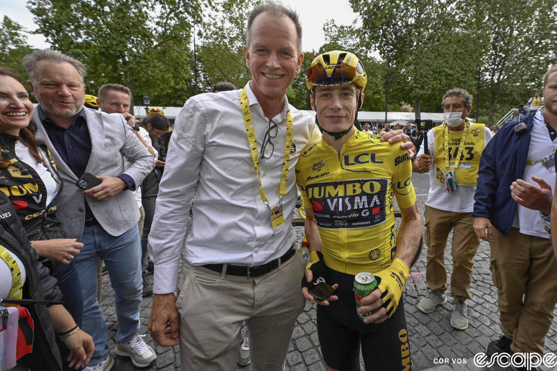 Jumbo-Visma general manager Richard Plugge stands with his arm around Jonas Vingegaard at the finish of the 2023 Tour de France in Paris. Vingegaard is still in team kit, including the yellow jersey of race winner, while Plugge is wearing the unremarkable outfit of khaki pants and a white long-sleeve button down shirt. Both are smiling, Plugge a bit more widely than Vingegaard.