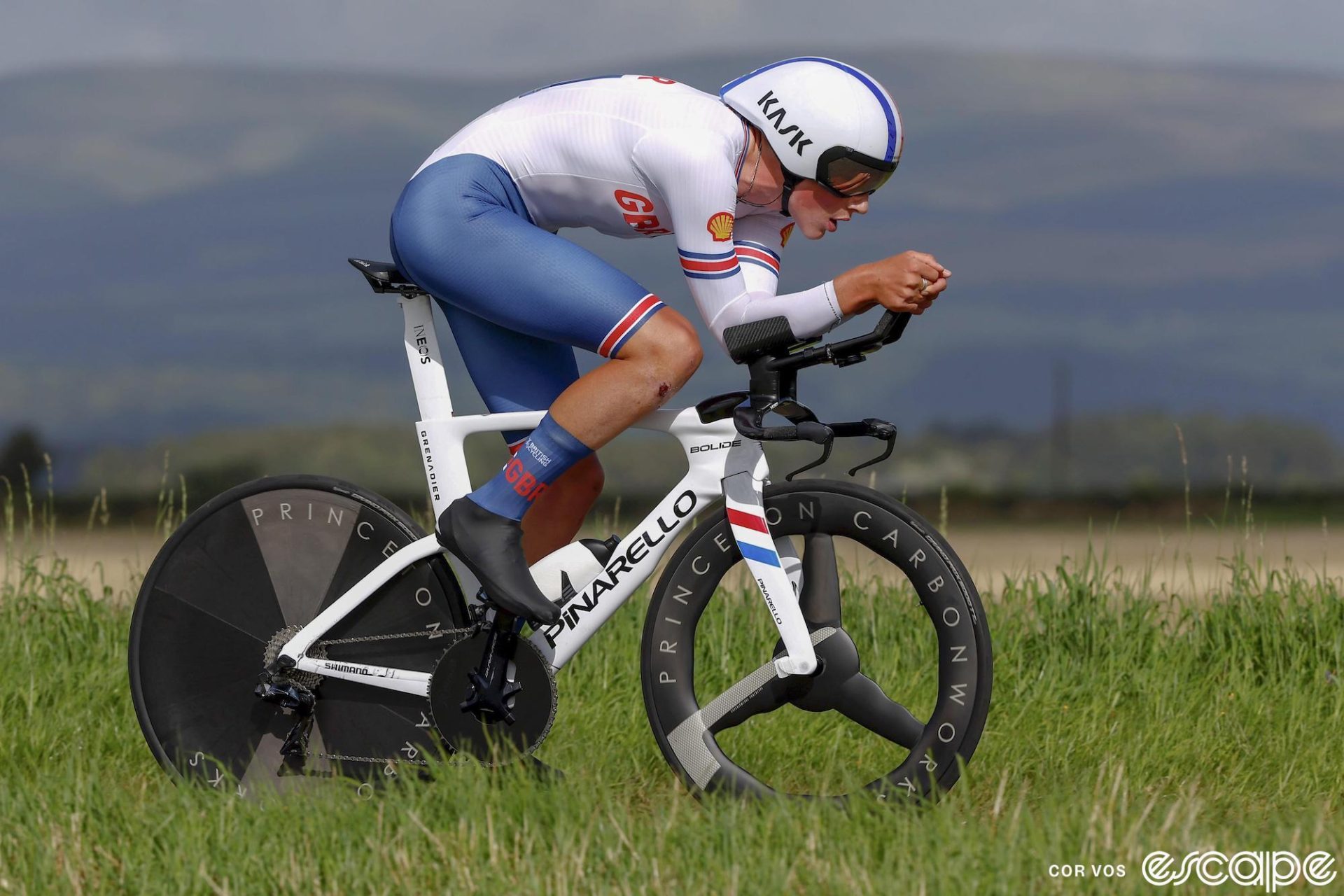 Britain's Joshua Tarling races across the Scottish landscape in the 2023 World Time Trial Championships. He's wearing the white and blue kit of the British national team, aboard a white Pinarello Bolide time trial bike, in an efficient aero tuck.
