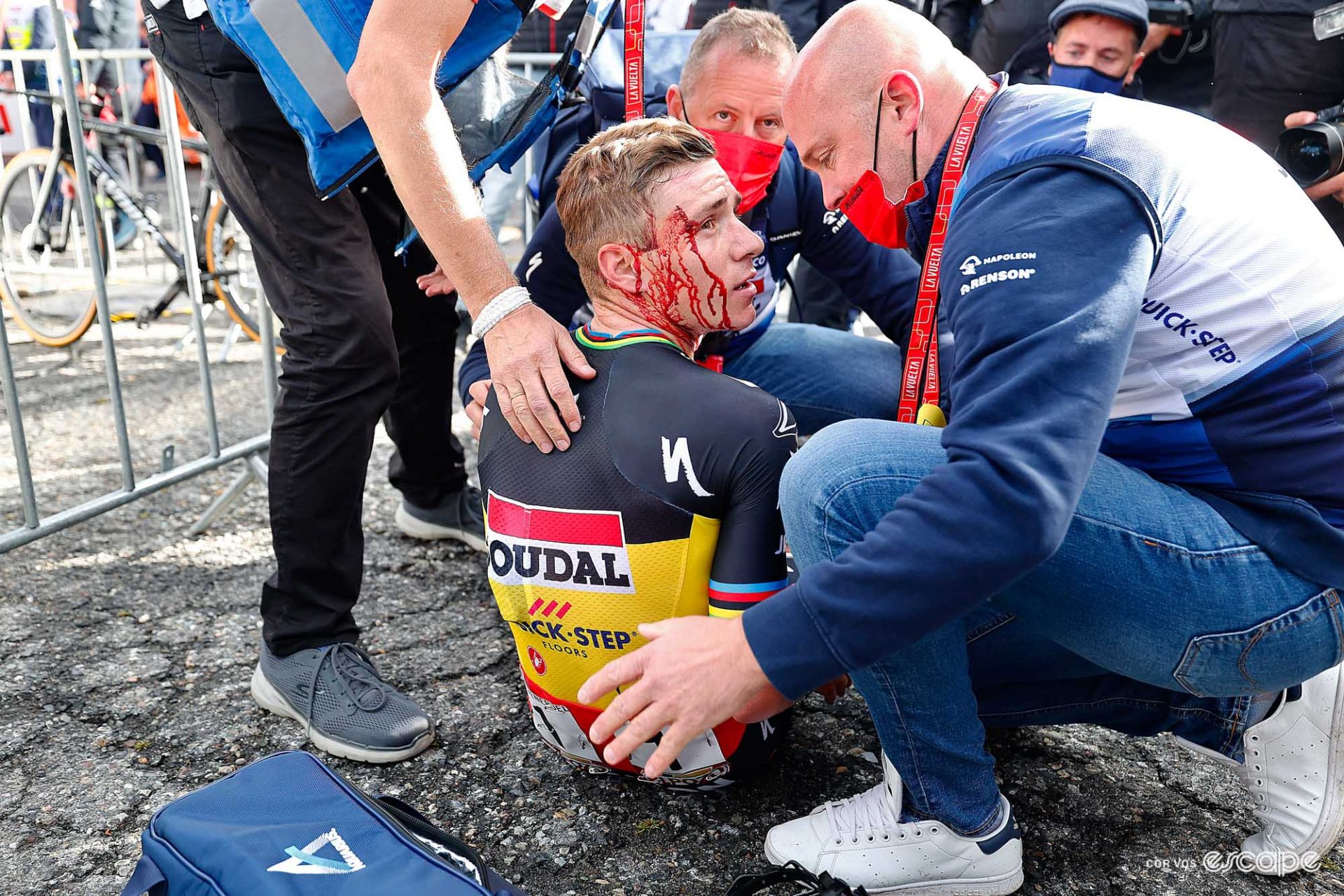 Evenepoel sitting on the ground at the end of stage 3, surrounded by team staff. He has blood all down the side of his face after crashing. 