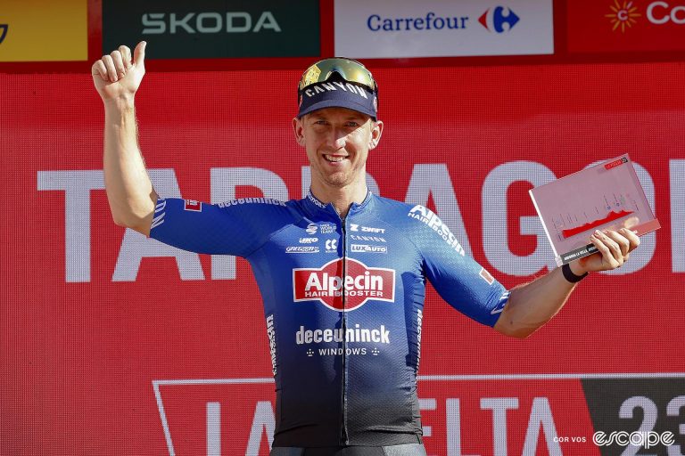 Kaden Groves stands on the presentation podium holding his right hand up in the air while his left hand holds a trophy for winning a stage of the Vuelta a España.