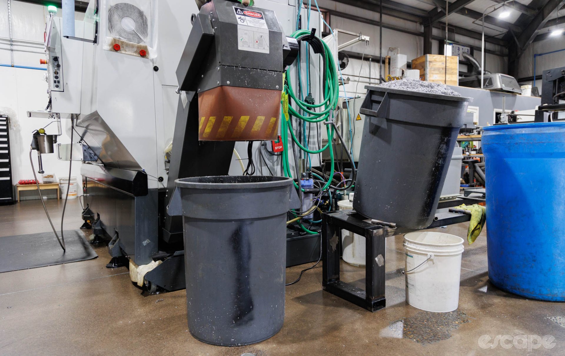 Two large trash cans sit next to the CNC mill. One is piled high with aluminum shavings. The other sits below a chute to catch more.
